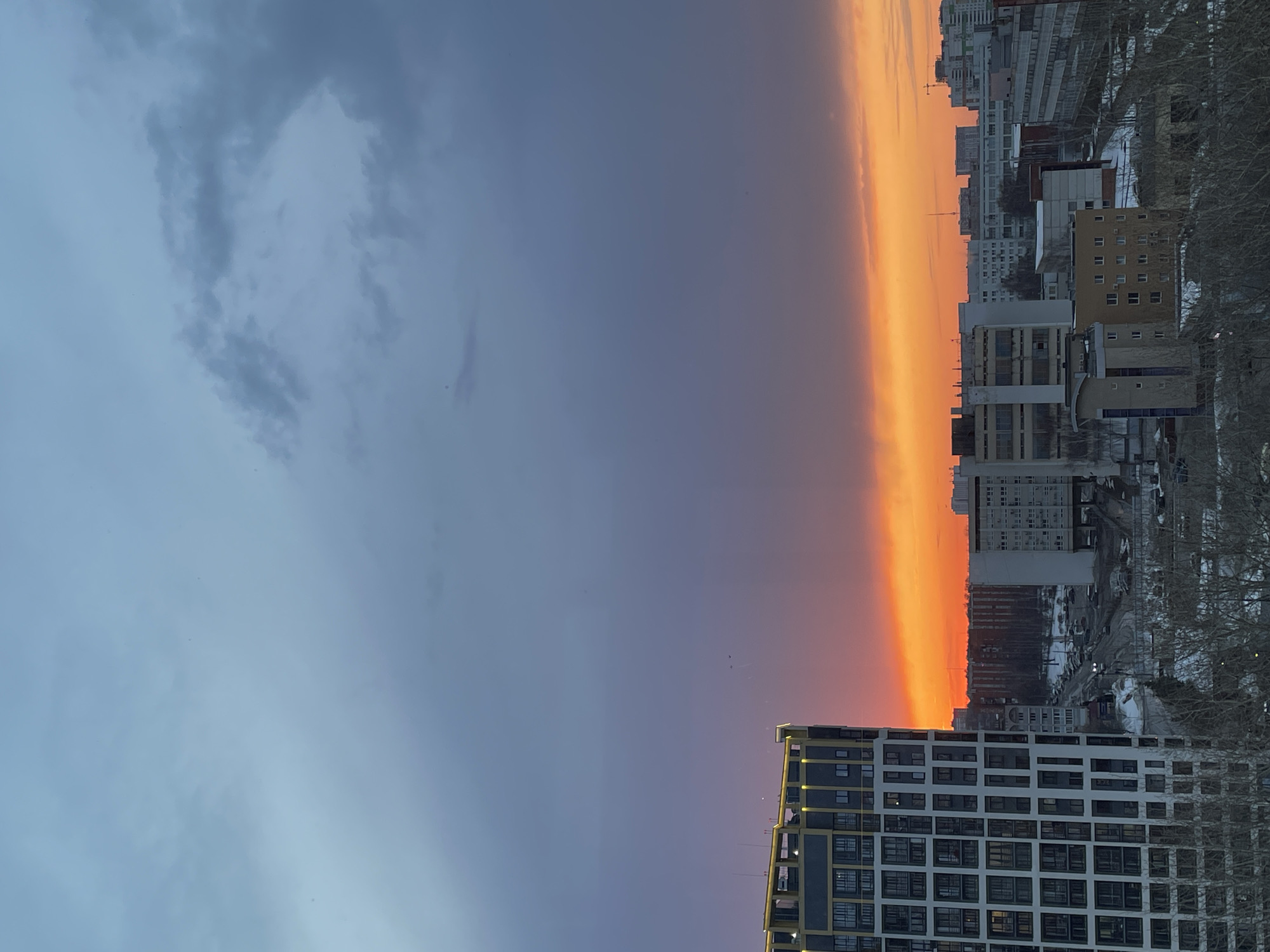The image shows a vertical view of a cityscape at sunset in Izhevsk, Russia. The sky is divided: the left side is filled with dark grey clouds tinged with blue and purple, and the right side glows with a vibrant orange and yellow hue as the sun sets. The right side of the image is dominated by buildings, including tall apartment blocks, commercial buildings, and residential structures. The facades are mostly white, grey, or brown, and windows are visible throughout. Trees with bare branches are also visible among the buildings, suggesting a cold season. Some snow patches are seen on the ground, adding to the winter ambiance. A modern high-rise building with a geometric window pattern dominates the bottom right corner.