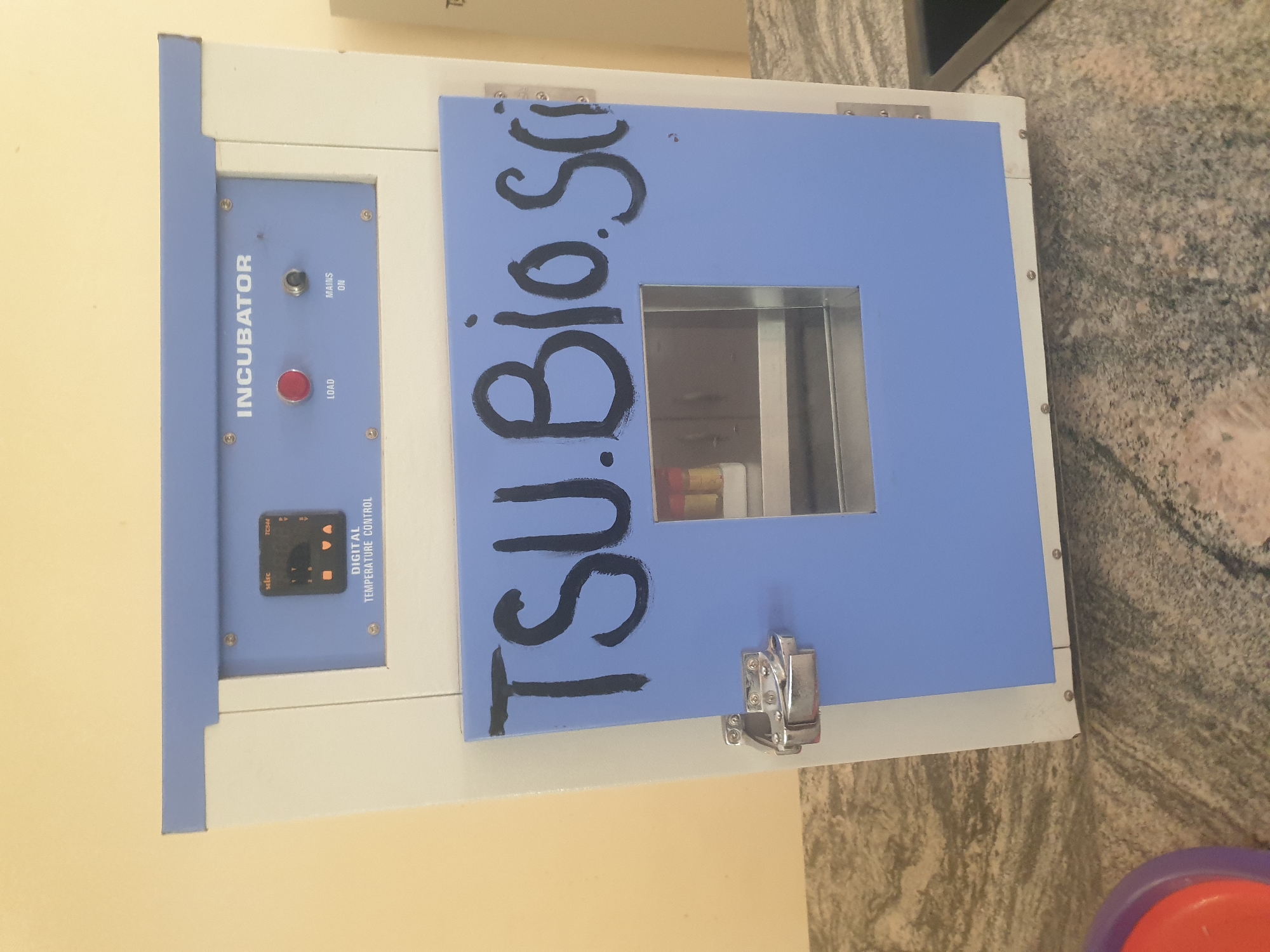 An incubator, labeled "INCUBATOR", is situated on a speckled gray countertop. The incubator is white with a light blue door. A black, hand-painted label on the door reads "TSU. Bio. Sci". A small rectangular window on the door reveals two small, capped vials inside. To the left of the window, a silver hinge is visible. The top of the incubator has a control panel with a digital temperature display, a red "LOAD" button, and a "MAINS ON" switch. The surrounding environment is dimly lit, with a plain yellow wall in the background. The overall scene is static and does not depict any individuals or specific actions. The provided context for the city of Jalingo, Nigeria is not reflected in the visual elements of the image itself.