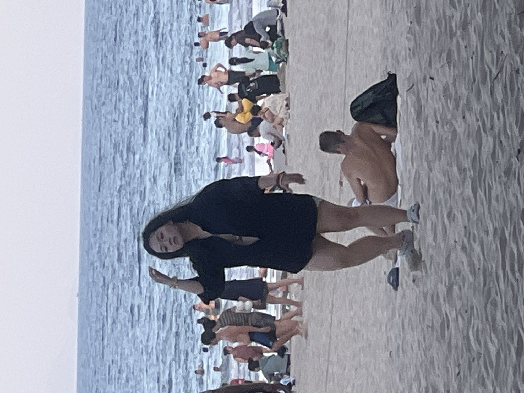 This is a photograph of a beach scene in Đà Nẵng, Vietnam, likely taken during daylight hours. The water is visible in the background with several people swimming or wading. The sand in the foreground is lightly textured with some scattered debris.
In the middle ground, a woman with long dark hair is standing on the sand, facing the camera. She is wearing a dark, unbuttoned shirt over what appears to be a bikini top, and light-colored shorts. Her right hand is raised to her hair. To her left, several other individuals are present on the beach, some walking and others sitting or standing. A man in the lower right corner of the frame is lying on his back on a towel, with a dark backpack next to him. The scene depicts a typical beach environment with various individuals engaged in leisure activities.