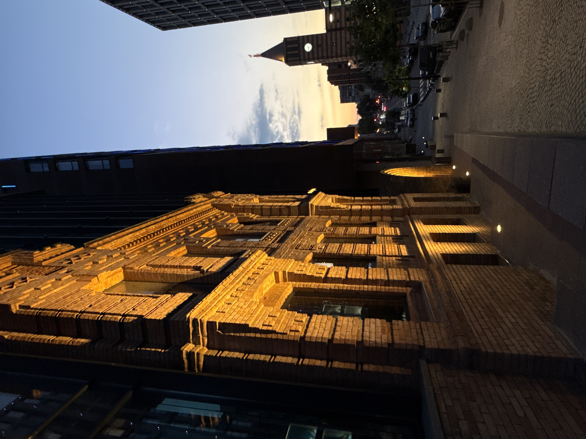 The image captures a street view in Dnipro, Ukraine, at dusk or early dawn. A historic brick building with illuminated architectural details stands on the left, contrasting with a modern, dark-colored skyscraper to its right. The street, paved with cobblestones, slopes downwards and is lined with streetlights and parked cars. In the distance, a clock tower with red and dark stripes is visible against a twilight sky painted with hues of orange, yellow, and blue. The scene depicts an urban environment transitioning from day to night.