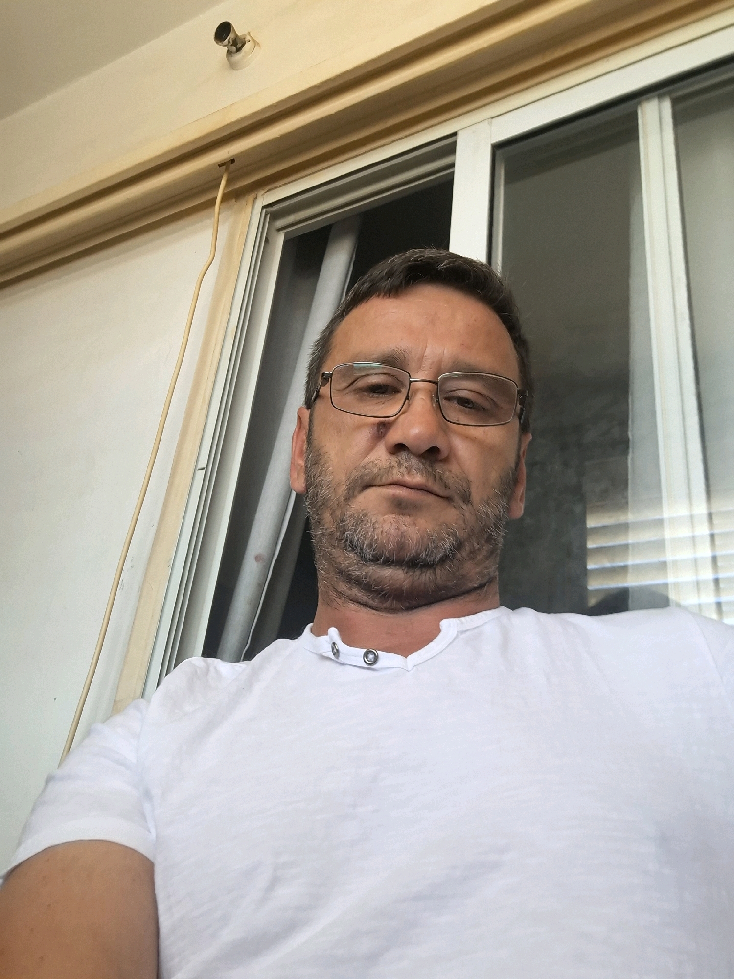 A man with short brown hair, a beard, and glasses is captured in a close-up, waist-up, selfie-style shot, likely taken indoors in Yerocham, Israel. He is wearing a white, short-sleeved t-shirt. Behind him, a sliding glass door with horizontal blinds is visible, suggesting he may be in a home or office setting. To the left of the frame, a light-colored wall and a wooden trim are present. The lighting suggests it is daytime.