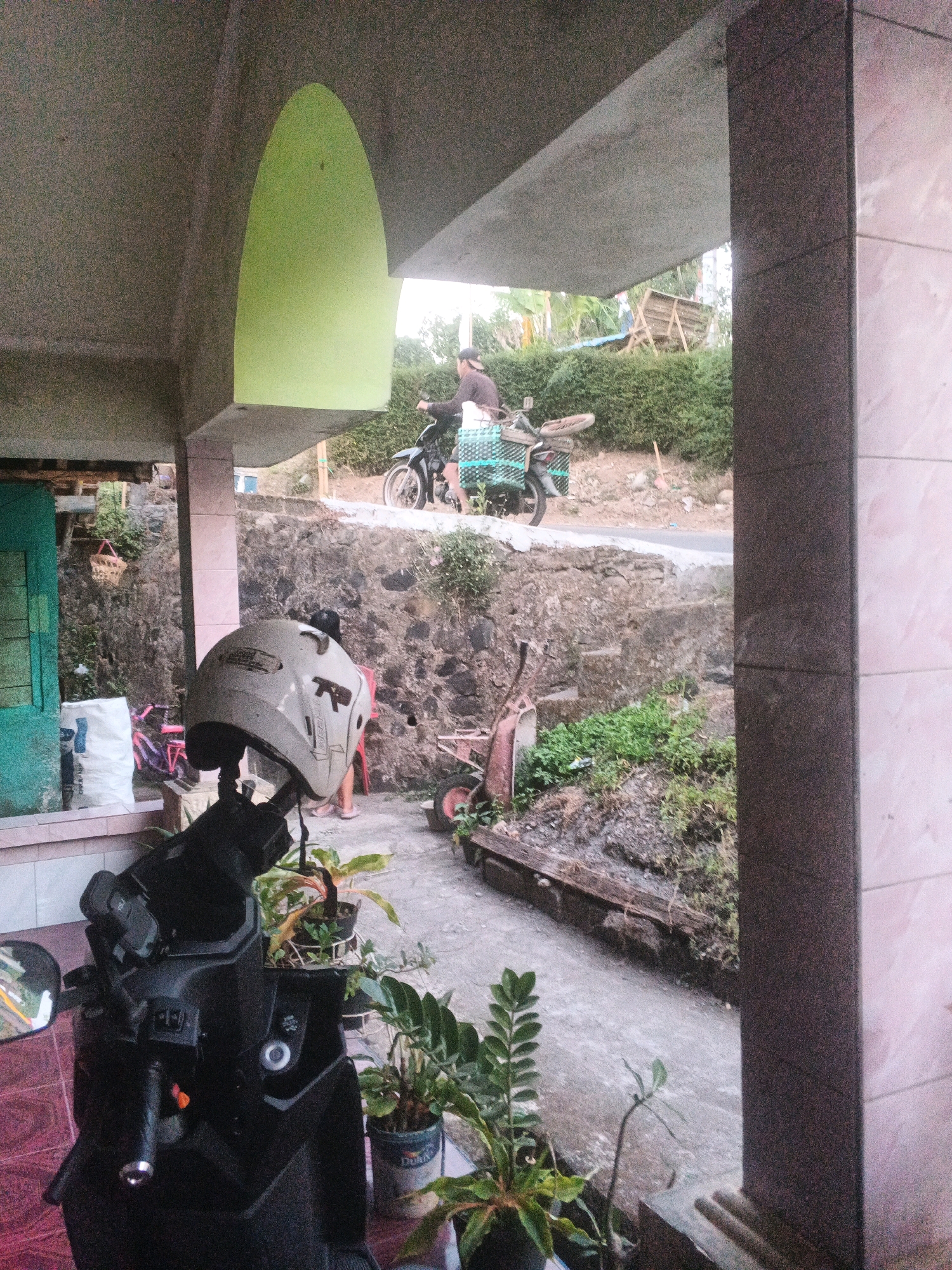 In Keji, Indonesia, a scooter is parked in front of a building. The scooter is black and has a white helmet with some writing on it resting on its handlebars. To the right of the scooter, a person is riding a motorcycle on a dirt road. The motorcycle is carrying a large green basket. The background consists of a stone wall, lush green bushes, and trees. The building has a concrete roof and a green wall section. A small cart with red wheels is also visible near the motorcycle.