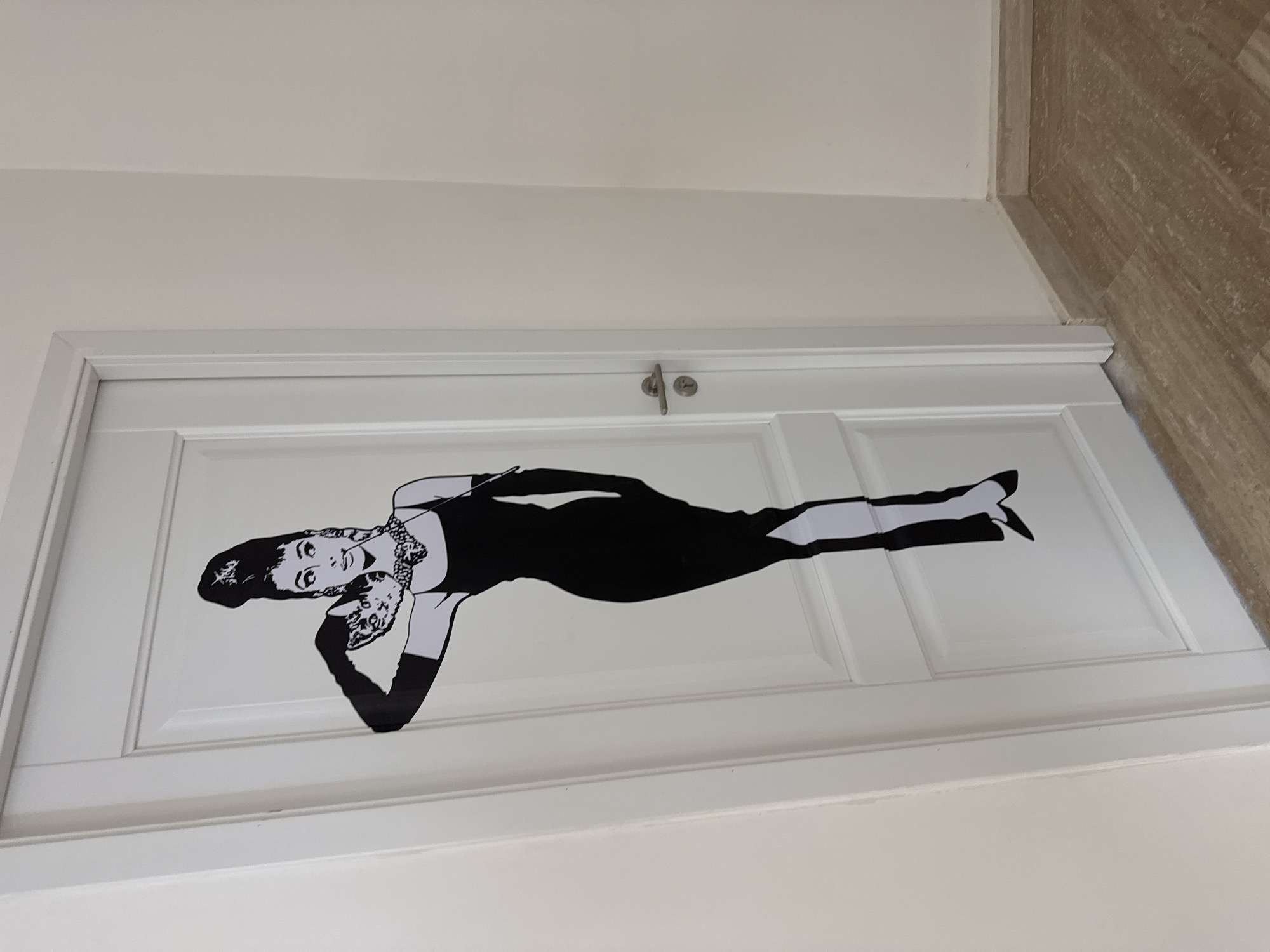 An indoor scene in Yerevan, Armenia, displays a white paneled door adorned with a large, black-and-white graphic of a woman, strikingly resembling Audrey Hepburn from "Breakfast at Tiffany's." The graphic depicts the woman in a classic pose, wearing a black sheath dress, long black gloves, and an elaborate necklace, with her hair styled in an updo. She appears to be lying down or leaning back gracefully, looking towards the viewer.
The door itself is a standard interior design, featuring recessed rectangular panels and a silver-colored door handle or lock mechanism. A small, round plate next to the handle has the word "Joud" embossed on it. The door is set within a light-colored, possibly off-white or cream, wall. To the right of the door and wall, a light brown floor with a wood-grain or tile pattern is visible, contributing to the clean and modern aesthetic of the space. The lighting is even, suggesting typical indoor illumination. There are no other people or distinct activities depicted; the focus is solely on the decorative door.