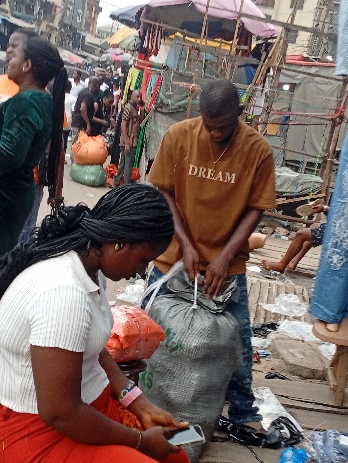 A busy outdoor marketplace scene shows a woman in a white shirt and red pants using a cell phone. Nearby, a man wearing a brown "DREAM" t-shirt ties a large gray sack. Other people stroll around, and fabric hangs from makeshift stalls. Litter and bags are scattered on the ground, with glimpses of buildings in the background. The scene is bustling with activity.