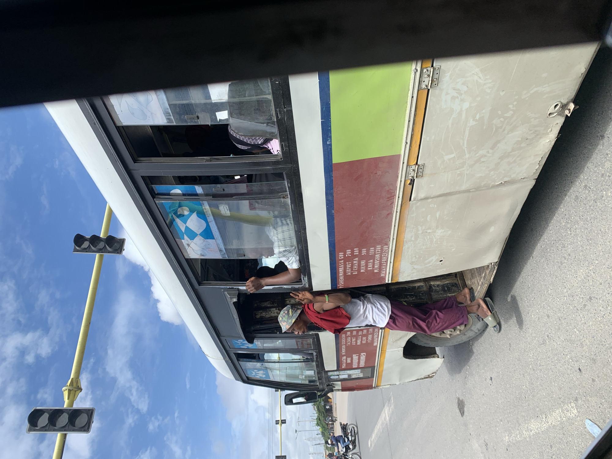 The image shows a man riding on the outside of a public transport bus in Dar es-Salaam, Tanzania. He is sitting on the wheel arch with his feet resting on the top of the tire, holding onto the bus window frame. He is wearing a hat, t-shirt, maroon-colored pants, and sandals. The bus is painted in shades of white, green, and maroon and has several route and fare details written on its side. In the background, there is a traffic light and other buses. The sky is visible with clouds and the image is taken from the inside of another vehicle.