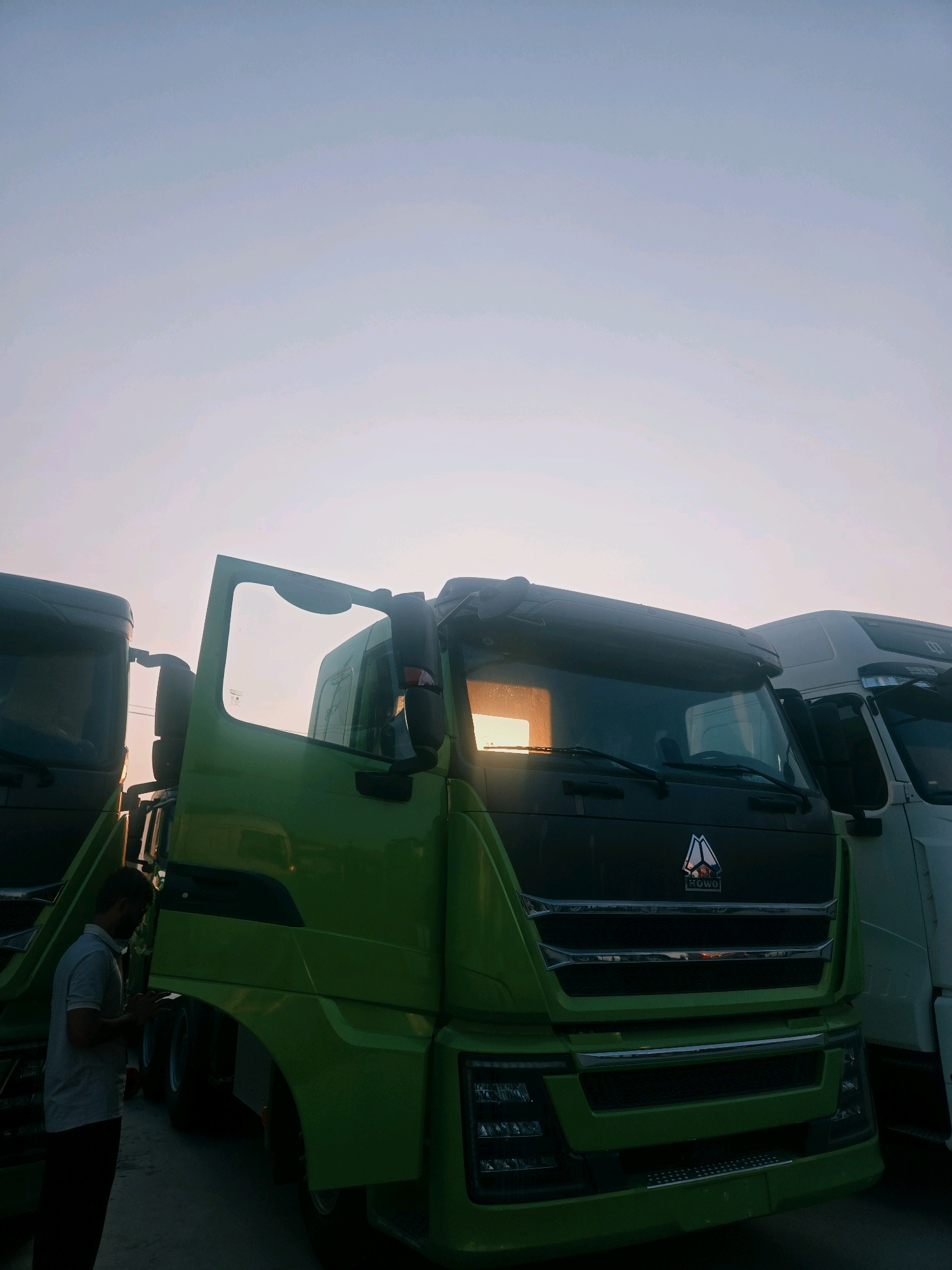 This is an outdoor, medium shot of a line of trucks parked in Liangshan County, China, during sunset. The sky is a pale blue with hints of pink, suggesting the sun is low on the horizon. In the foreground, a bright green truck dominates the frame, its driver's side door open. A man in a light grey shirt and dark pants stands to the left of the green truck, looking down at his phone. His back is mostly to the camera, and he is partially obscured by the truck. The front of the green truck features a prominent chrome grille with a red and white logo. To the right of the green truck, part of a white truck is visible, also appearing to be a large commercial vehicle. The ground appears to be paved. The overall impression is of a truck yard at the end of the day.