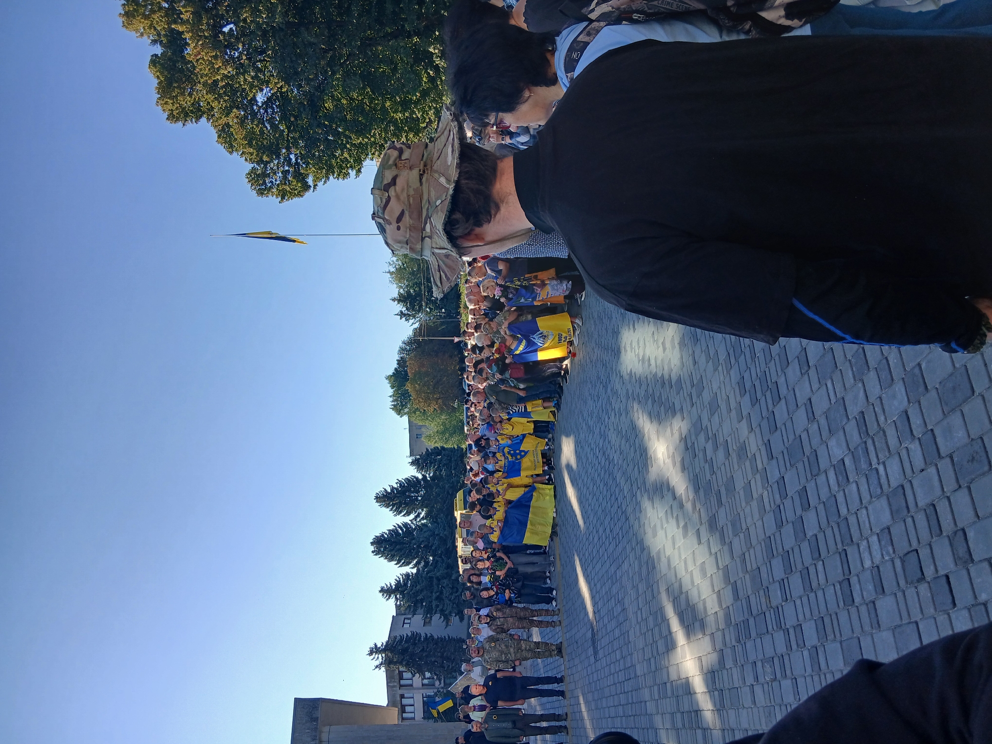 A crowd of people, some holding Ukrainian flags, are gathered in an outdoor paved area in front of a modern building with a concrete facade and a flagpole flying the Ukrainian flag under a clear blue sky. The crowd includes adults and children, with some individuals in military-style camouflage uniforms. The lighting suggests it is daytime, with shadows cast on the pavement by an unseen overhead structure or trees. The overall atmosphere appears to be solemn or commemorative. The location is identified as Verkhnodniprovsk, Ukraine.