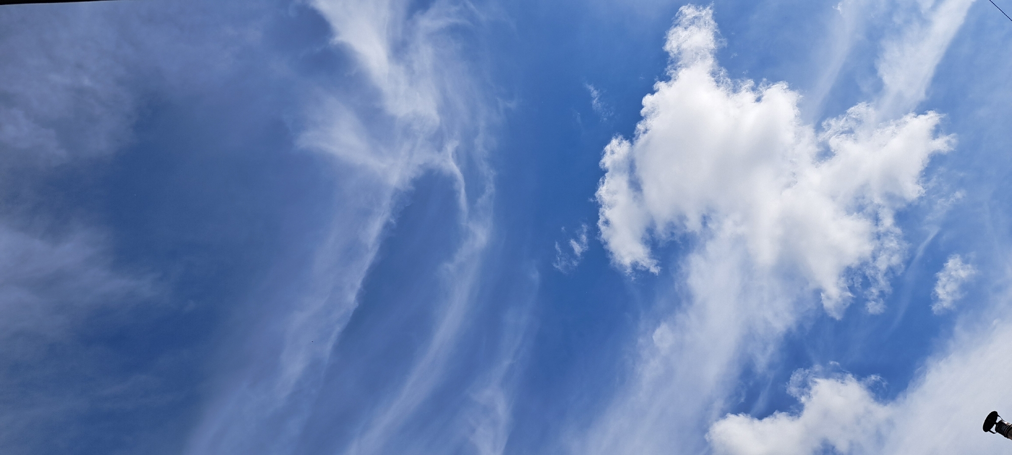 The image shows a clear blue sky with wispy white clouds. Some of the clouds are thin and streaky, while others are puffy and cumulus-like. There is a dark cylindrical object with a black cap visible in the bottom left corner of the image, possibly a chimney or vent pipe. The scene is an outdoor environment, likely during the daytime, given the bright sky. No people or specific city landmarks are identifiable, so its location in Азьмушкино, Russia cannot be confirmed from the visual information alone.