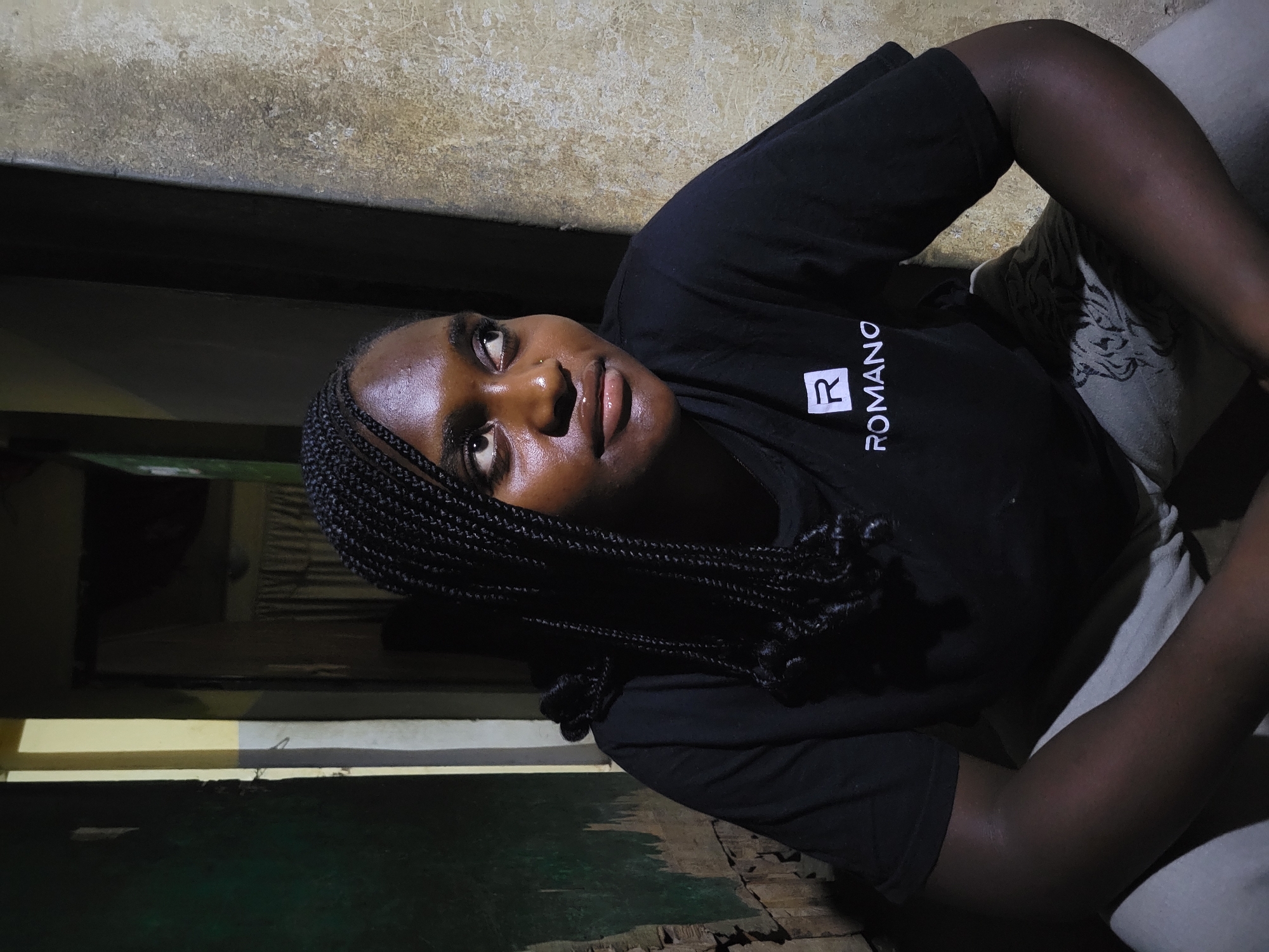 A medium shot of a woman with dark skin and braided hair is featured, wearing a black t-shirt with the word "ROMANO" on the left side. She is positioned indoors, angled upward toward the left. The background includes a wall with visible texture and paint, along with a dark wooden frame, and what appears to be a bed. Given the prompt's direction, it is assumed that the image was taken in Jos, Nigeria.