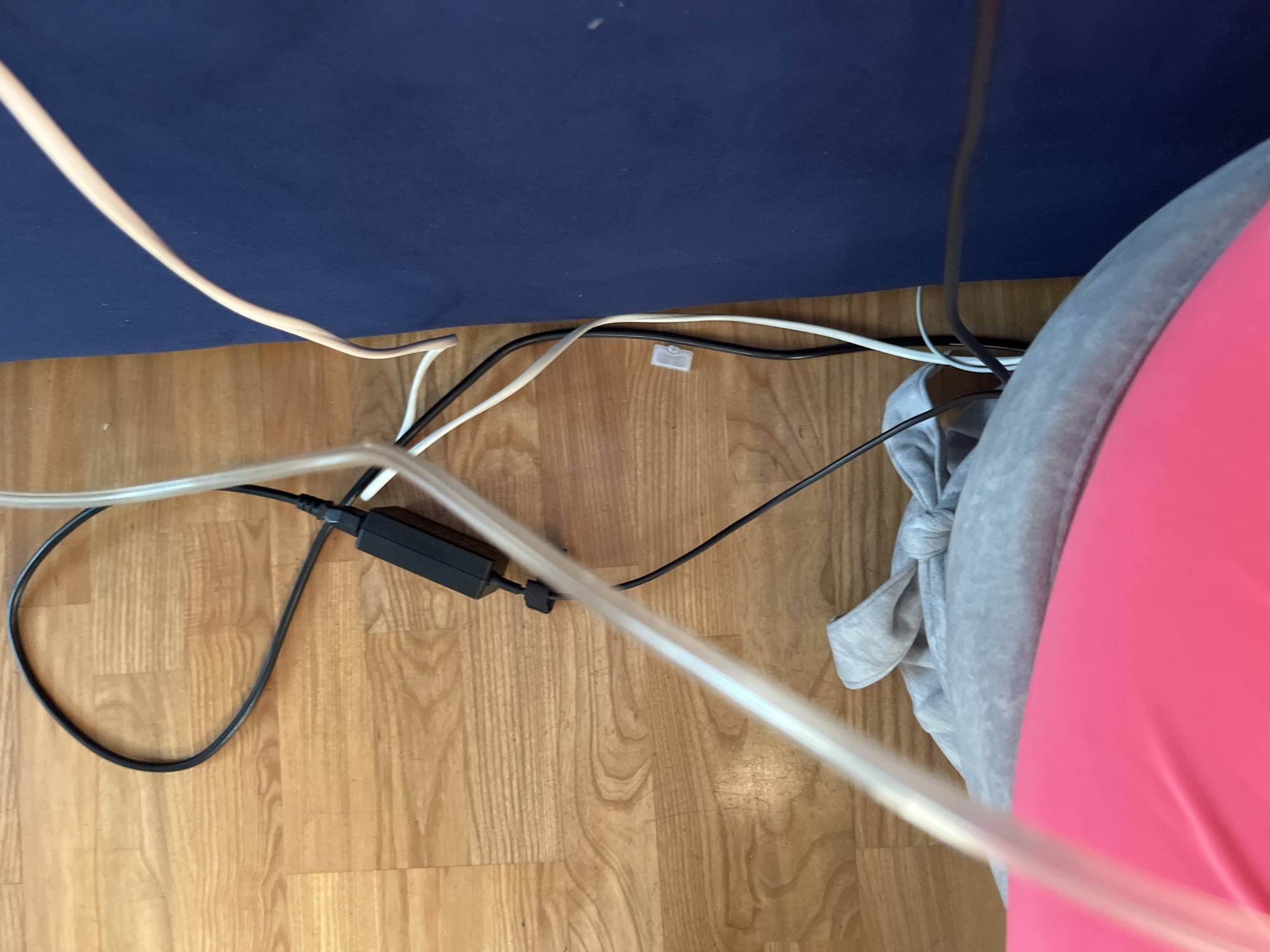 The image shows a close-up view of a wooden floor with various cables and a piece of gray fabric with a bow. A thick, clear tube extends diagonally from the bottom left of the frame upwards. To the right of the tube, a gray fabric item, possibly a garment or blanket, is visible, with a prominent bow tie shape.
Further to the right, several cables are present: a thick black cable with a rectangular black adapter, a thin white cable, and another thin black cable. These cables are connected to an unseen power source. A dark blue surface, likely a piece of furniture, occupies the right side of the image.
The scene is indoors, on a wooden floor. The lighting suggests an overhead light source. There are no people or animals visible in the image, nor any clear indication of the specific location within Saratov, Russia. The perspective is from a high angle, looking down.