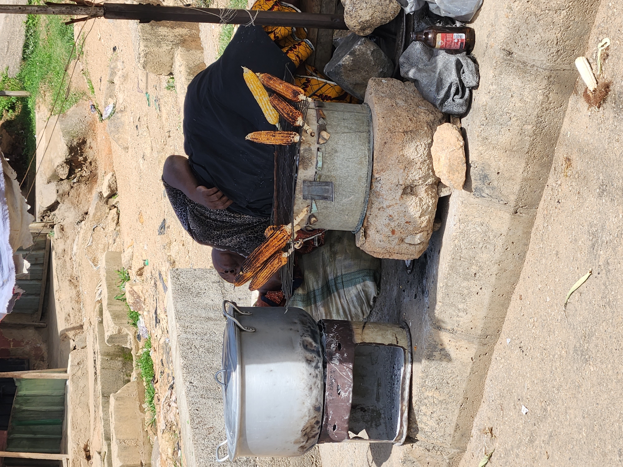 A street vendor in Kabong, Nigeria, grills corn on a makeshift stove constructed from a metal drum and rocks. The vendor, draped in a dark shawl over patterned clothing, sits beside the stove, overseeing the roasting corn. A large metal pot sits on another cooking surface nearby. A mix of trash and debris surrounds the cooking area. A building's steps and facade are visible behind the vendor. A bottle and gray bag sit on top of a pile of rocks near the stove. The ground is dry and dusty.