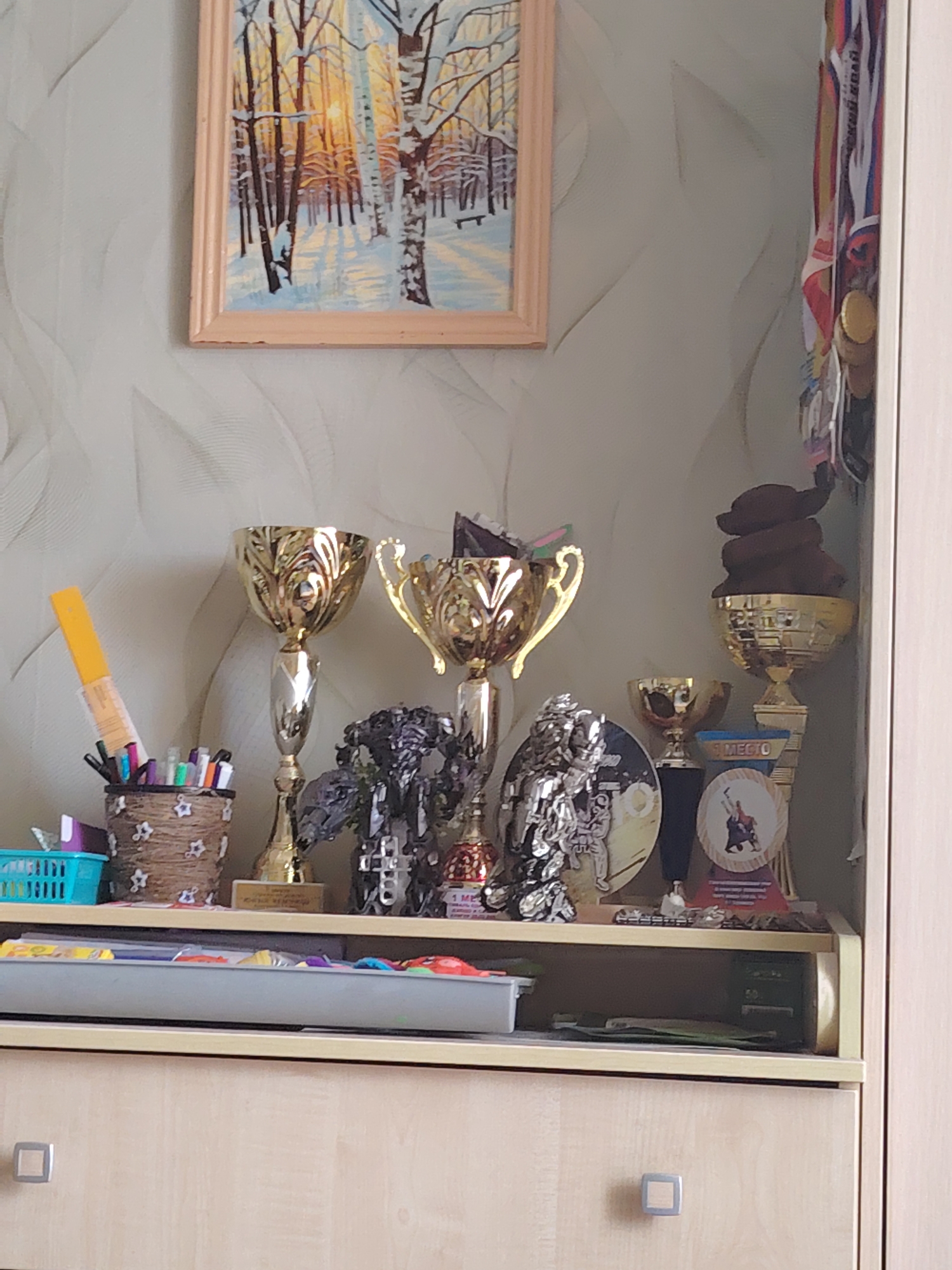 The image displays a shelf or desk with various decorative items and supplies, possibly in a home environment in Chaikovsky, Russia. On the upper shelf, a framed painting of a winter forest scene with birch trees and sunlight is hung. To the right of the painting, a collection of medals with ribbons is attached to the wall.
The main focus of the image is on the shelf itself. A variety of trophies are displayed, including a tall, ornate golden trophy on the left, another similar golden trophy to its right, and a smaller golden trophy further to the right, topped with a dark, abstract object. There are also some metallic figurines and circular decorative items, including one with a depiction of skaters and text. A blue plastic basket containing some items is visible on the far left. In front of the trophies, a brown woven container filled with pens and markers stands. A yellow marker is sticking out of this container.
Below the upper shelf, a lower shelf or drawer front is visible. This surface holds a tray containing what appear to be art supplies or stationery items, with colorful edges. The lower part of the image shows the front of a light-colored wooden drawer with a square handle. The overall impression is of a decorated display area. The lighting suggests an indoor setting with ambient light.