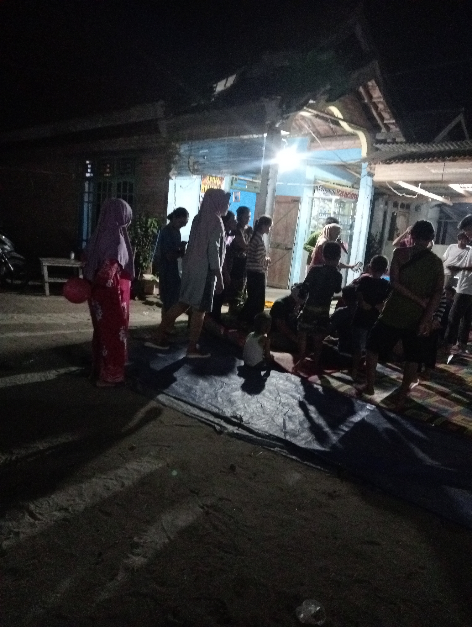The image depicts a nighttime gathering of approximately twelve people, a mix of adults and children, in what appears to be a residential outdoor area in Rasuan Baru, Indonesia. A blue and white building with a corrugated metal roof is visible in the background. Several individuals are standing and interacting, while some children are seated or positioned lower to the ground. A dark blue tarp is spread on the ground in the foreground, and a wooden table is set up to the left of the frame. The lighting suggests artificial illumination, likely from the building or portable lights. One person on the left is wearing a red and white patterned garment and a purple headscarf, holding a red balloon. Another individual in a purple headscarf and a light-colored outer garment is standing in the center-left. The overall atmosphere appears to be informal.