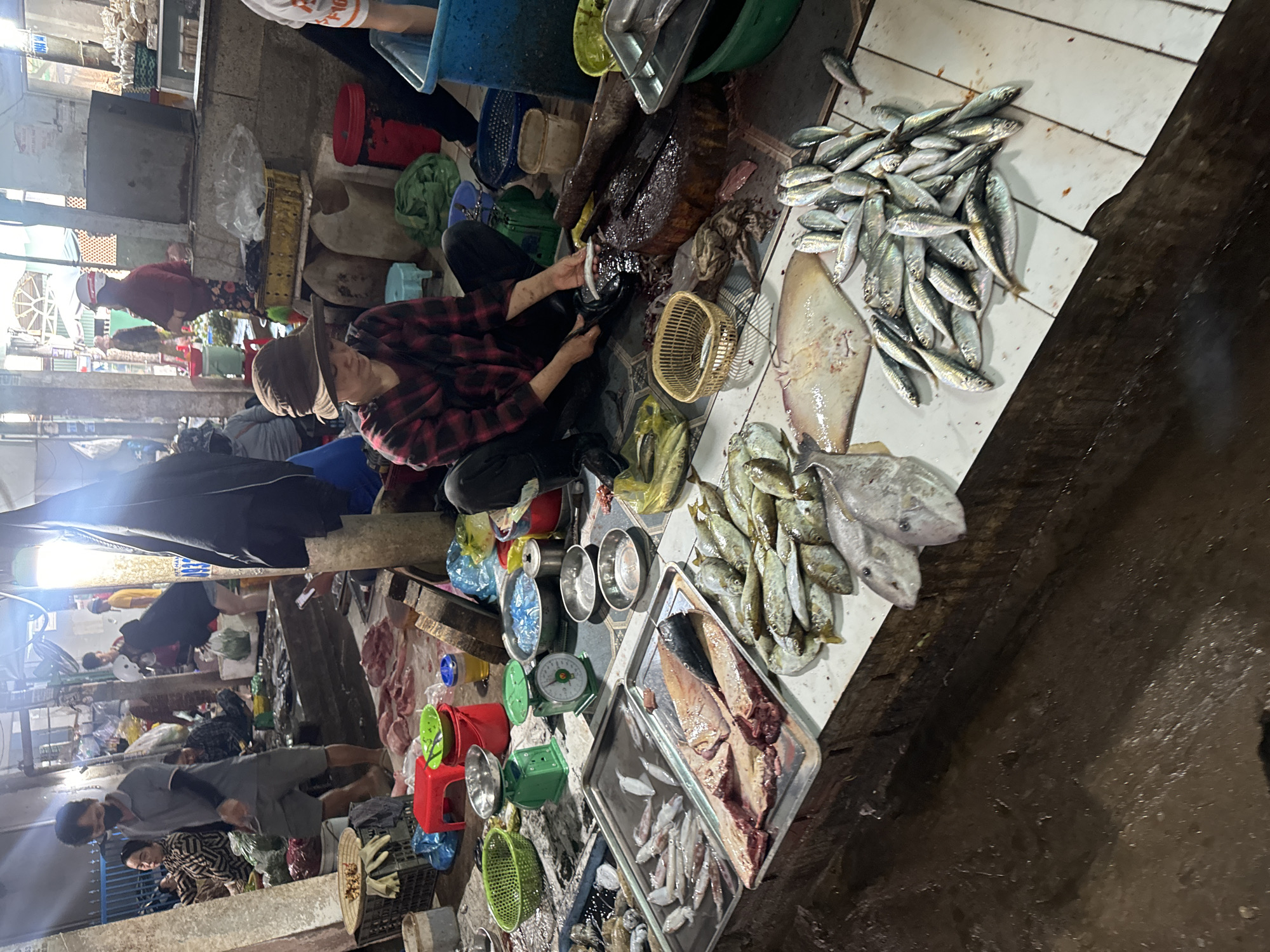 The image depicts a bustling fish market scene in Mũi Né, Vietnam. A woman wearing a wide-brimmed hat and a plaid shirt is seen seated on the floor, actively preparing fish with a knife on a large wooden chopping block. She is surrounded by a variety of seafood, including several trays of raw fish and a large stingray laid out on a white surface. Other individuals are visible in the background, engaged in market activities, with some appearing to be vendors or shoppers. The market environment is characterized by its open-air setup, with a combination of stalls, displays of fresh produce, and various containers, including buckets and baskets. The ground appears wet and somewhat muddy, indicating a typical market condition. The overall atmosphere suggests a vibrant and active commercial hub for fresh seafood.