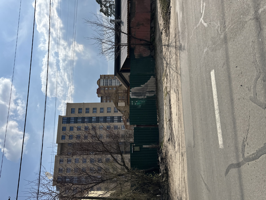 The image shows a street in Horenka, Ukraine (latitude: 50.525, longitude: 30.241) with a paved asphalt road in the foreground. The road is flanked by a sandy shoulder on each side. A green metal fence runs along the shoulder on the right side of the image. Behind the fence is a partially destroyed building with a red metal roof, a beige building, and a green tree on the left. The sky is partially cloudy. There is a white painted lane marker on the road. There are no people visible in the image. The scene appears peaceful and abandoned.