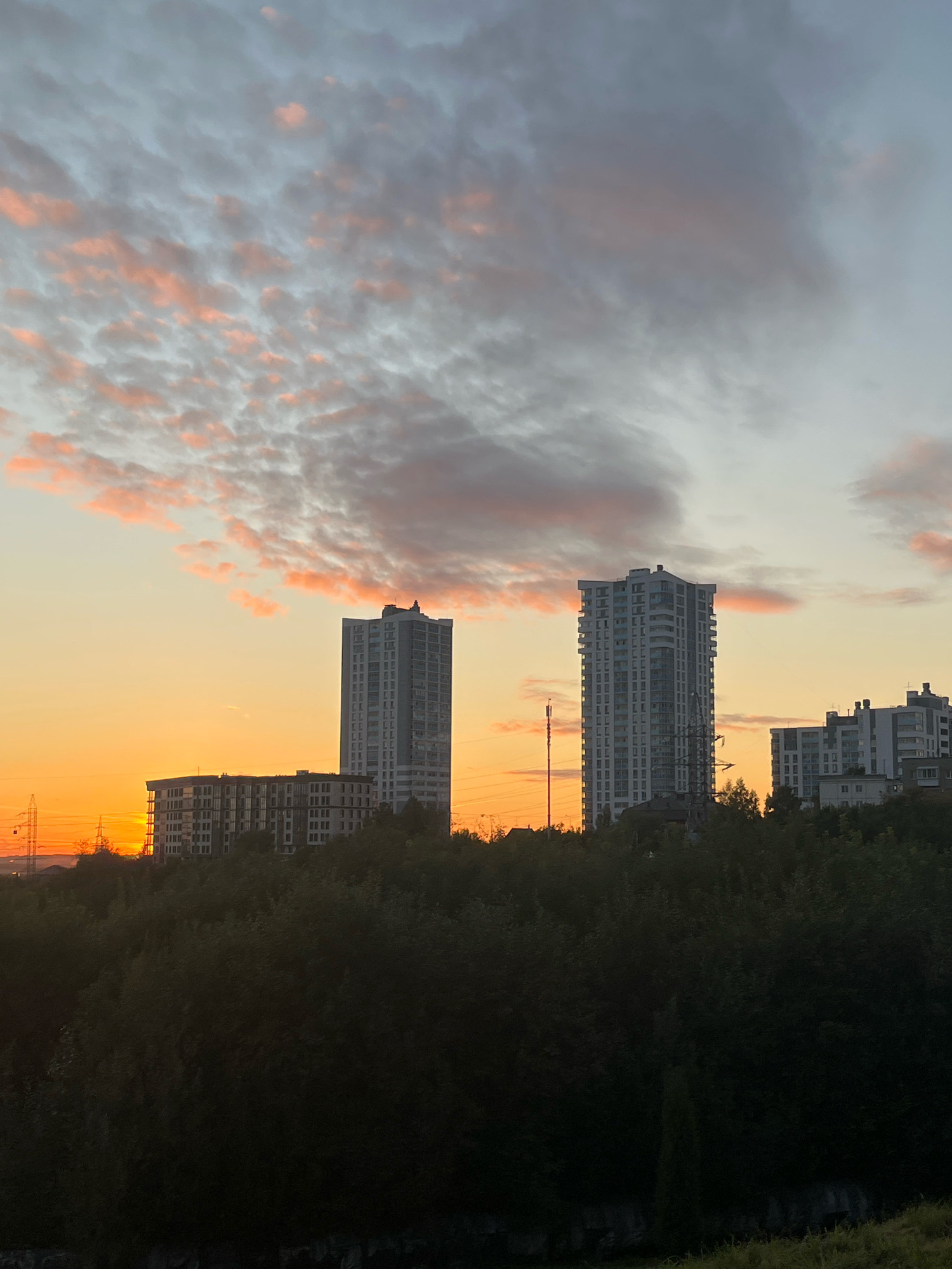 The image shows a sunset scene in Perm, Russia, with modern apartment buildings visible on a hillside. The sky is filled with a gradient of orange and pink hues, with scattered clouds. The buildings are tall and contemporary, with numerous windows. In the foreground, there is a dense area of dark green trees. Power lines and towers are also visible on the left side of the image, indicating an urban or suburban setting. There are no people or animals depicted in the image. The overall atmosphere is calm and serene, characteristic of a sunset in a city.