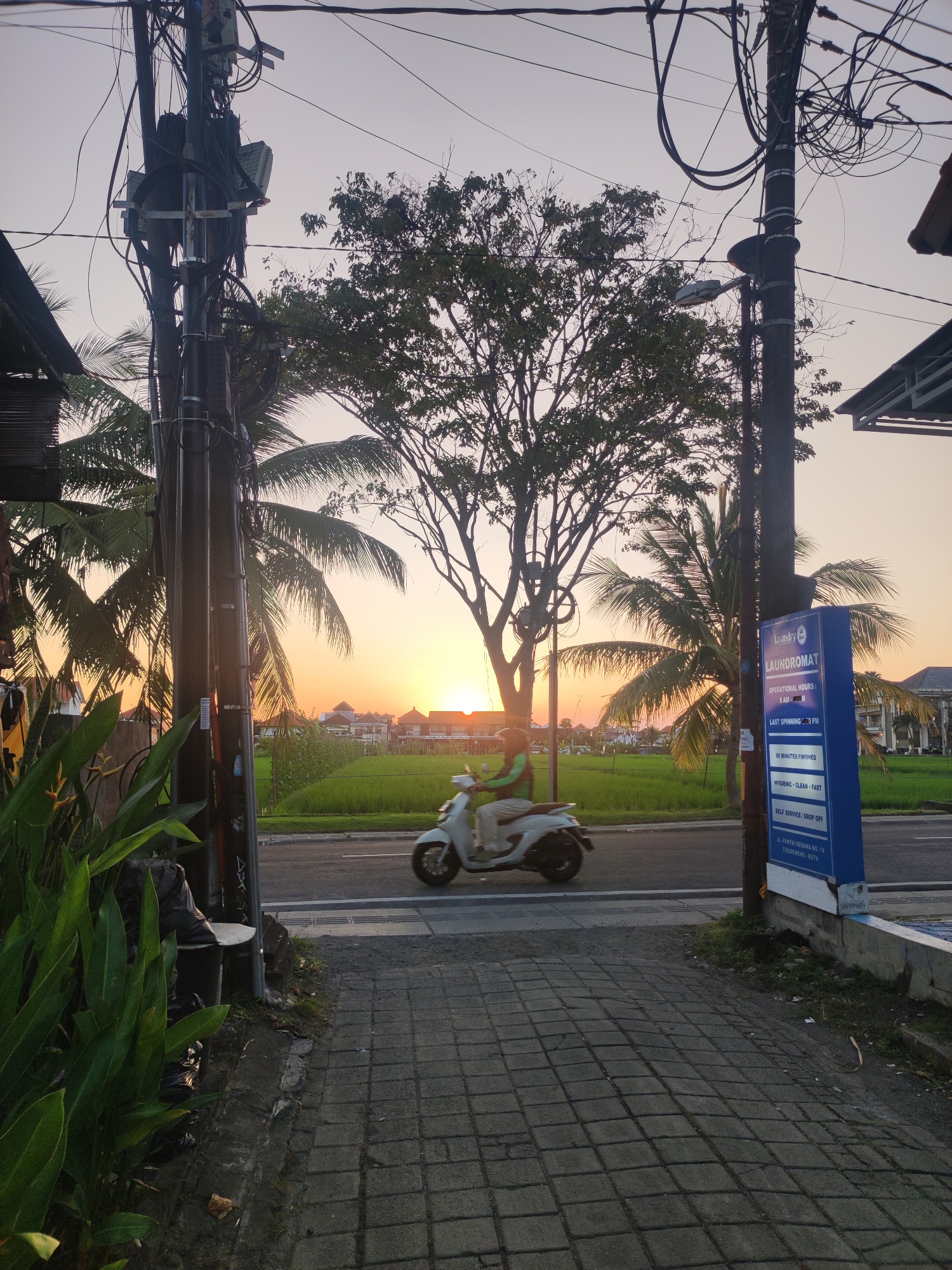 A street scene in North Kuta, Indonesia, at sunset. The view from a brick-paved sidewalk encompasses a street with a scooter in motion. Utility poles, entangled with wires, frame the scene. Palm trees and a large tree are visible against the setting sun, beyond a field of green. To the right, a blue Laundromat sign provides details including "JL. PANTAI BERAWA NO. 1X, TRUBENENG KUTA" and operational hours. A trash container is visible to the left, alongside flowering plants.