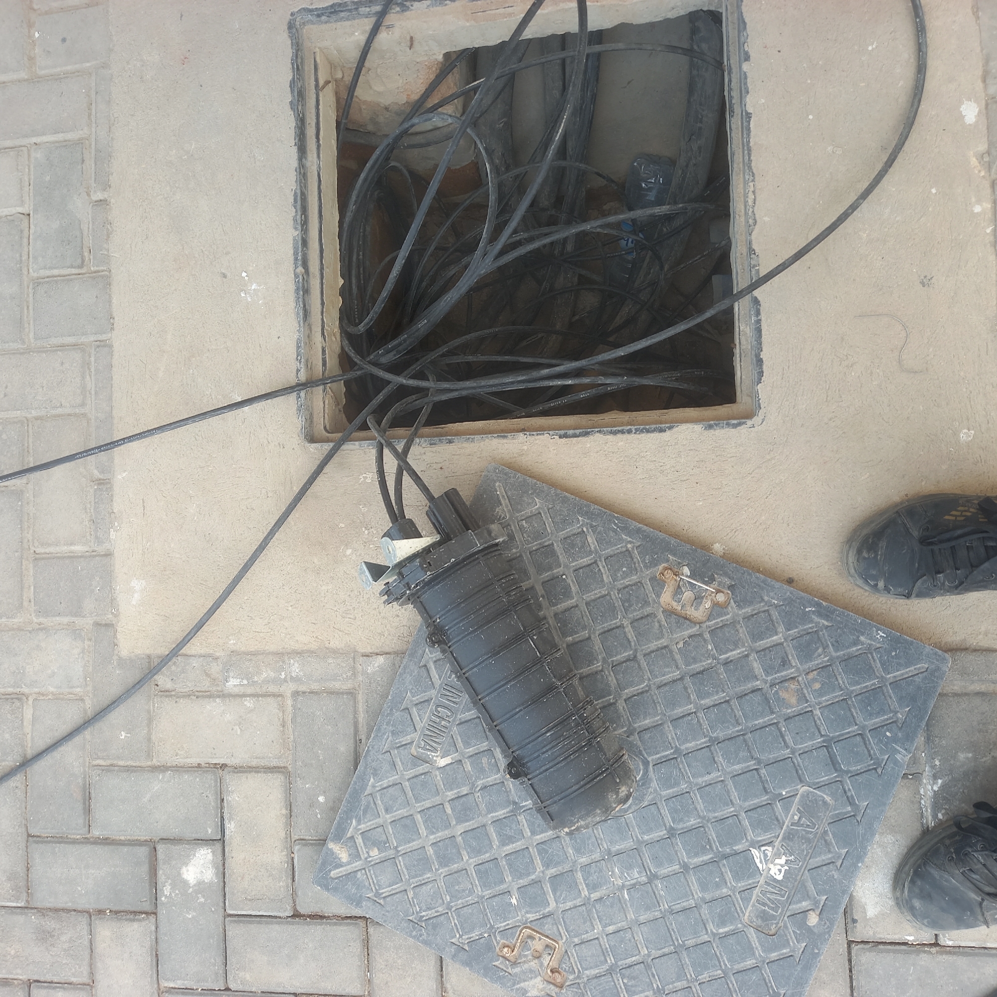 The image shows an open utility access point on a paved surface in Bububu, Tanzania. A large, black, rectangular utility cover is partially removed and lies on the ground next to the access point. Inside the open access point, a dense bundle of black cables is visible. A black cylindrical splice closure, a component used to join fiber optic cables, is resting on the utility cover. Two pairs of black shoes are visible at the bottom of the frame, suggesting people are present at the scene, likely involved in the utility work. The environment consists of interlocking grey pavers and a light-colored, sandy-textured concrete surface surrounding the access point. The overall impression is one of ongoing or recently completed infrastructure maintenance.