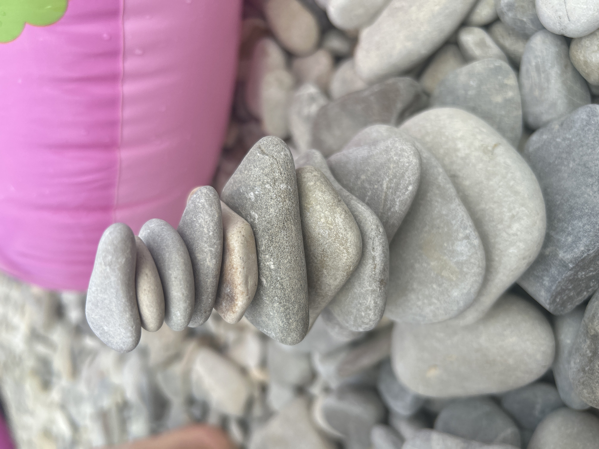 A rock cairn, a stack of smooth, grey, and beige pebbles, is the primary subject of the image, set against a blurred background of more pebbles and a large, bright pink inflatable object, possibly a pool float. The cairn is constructed with at least eight visible stones, progressively smaller towards the top, creating a balanced, vertical structure. The pebbles exhibit varied textures and colors, from pale grey to slightly darker grey, with some having subtle brown or beige undertones. The environment appears to be a beach or a shoreline, consistent with the presence of pebbles and an inflatable float. The lighting suggests an outdoor setting, likely during daytime. A portion of a person's arm and hand, with skin tone, is visible on the left side of the frame, suggesting human presence and interaction with the scene. The composition is close-up, focusing on the stacked rocks and the foreground pebble distribution. The location is identified as Gelendzhik, Russia.