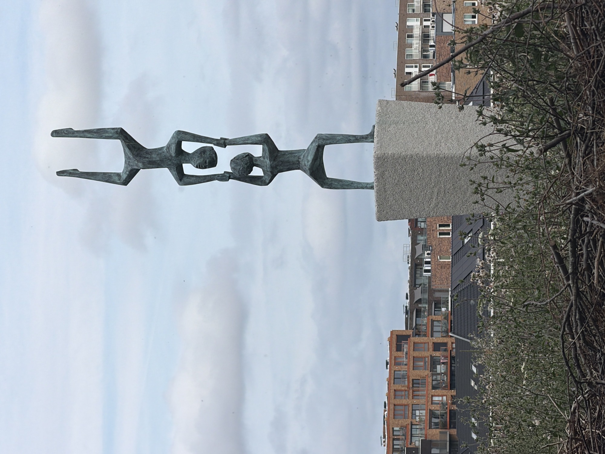 A bronze sculpture depicting three human figures in a stacked formation stands on a grey stone pedestal in Rijswijk, Netherlands. The bottom figure is kneeling, supporting the middle figure who is standing and holding up the top figure. The top figure is in a handstand position. The sculpture is situated in an urban environment with modern buildings in the background. In the foreground, a dense cluster of dry branches and green foliage is visible. The sky is overcast with scattered clouds.