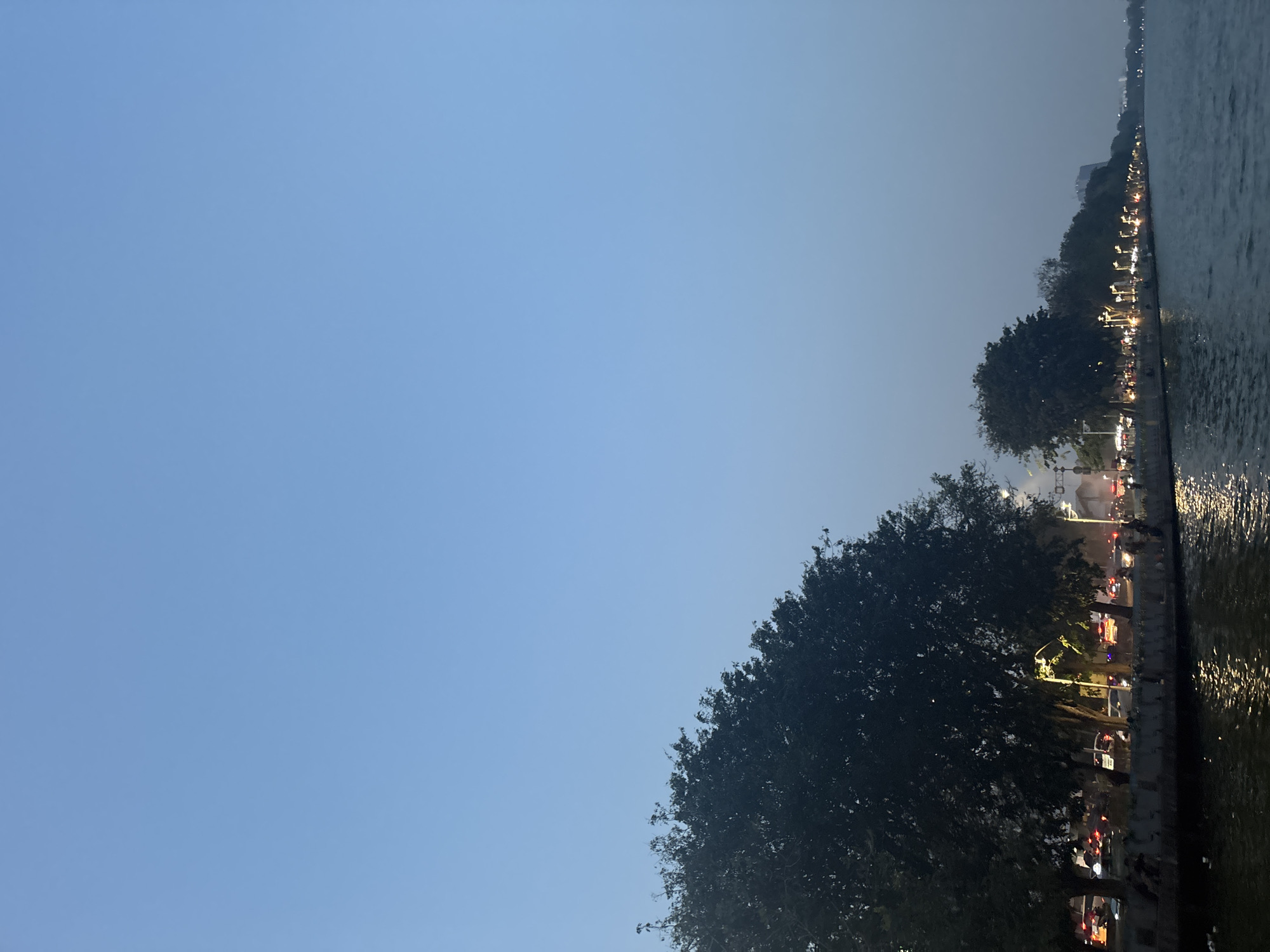The image depicts a waterfront scene in North Jakarta, Indonesia, likely at dusk. The sky is a clear, pale blue, transitioning to a lighter hue towards the horizon. On the left side of the frame, a large, dark green tree dominates the foreground, its foliage dense and casting shadows.
Beyond the tree, a street runs parallel to the waterfront, lined with streetlights that cast a warm glow. Several vehicles, visible as streaks of light and blurred shapes, are present on the street, suggesting moderate traffic. The street is bordered by a low wall, and beyond that, a row of smaller trees and some structures are visible. A few indistinct figures of people can be seen walking or sitting along the waterfront.
The foreground of the image features a body of water, likely a canal or bay, with its surface reflecting the lights from the street and sky, creating shimmering patterns. The overall atmosphere is serene, with the transition from day to night creating a peaceful ambiance. The scene appears to capture a moment of urban life by the water, characterized by the interplay of natural light, artificial illumination, and the calm movement of water.