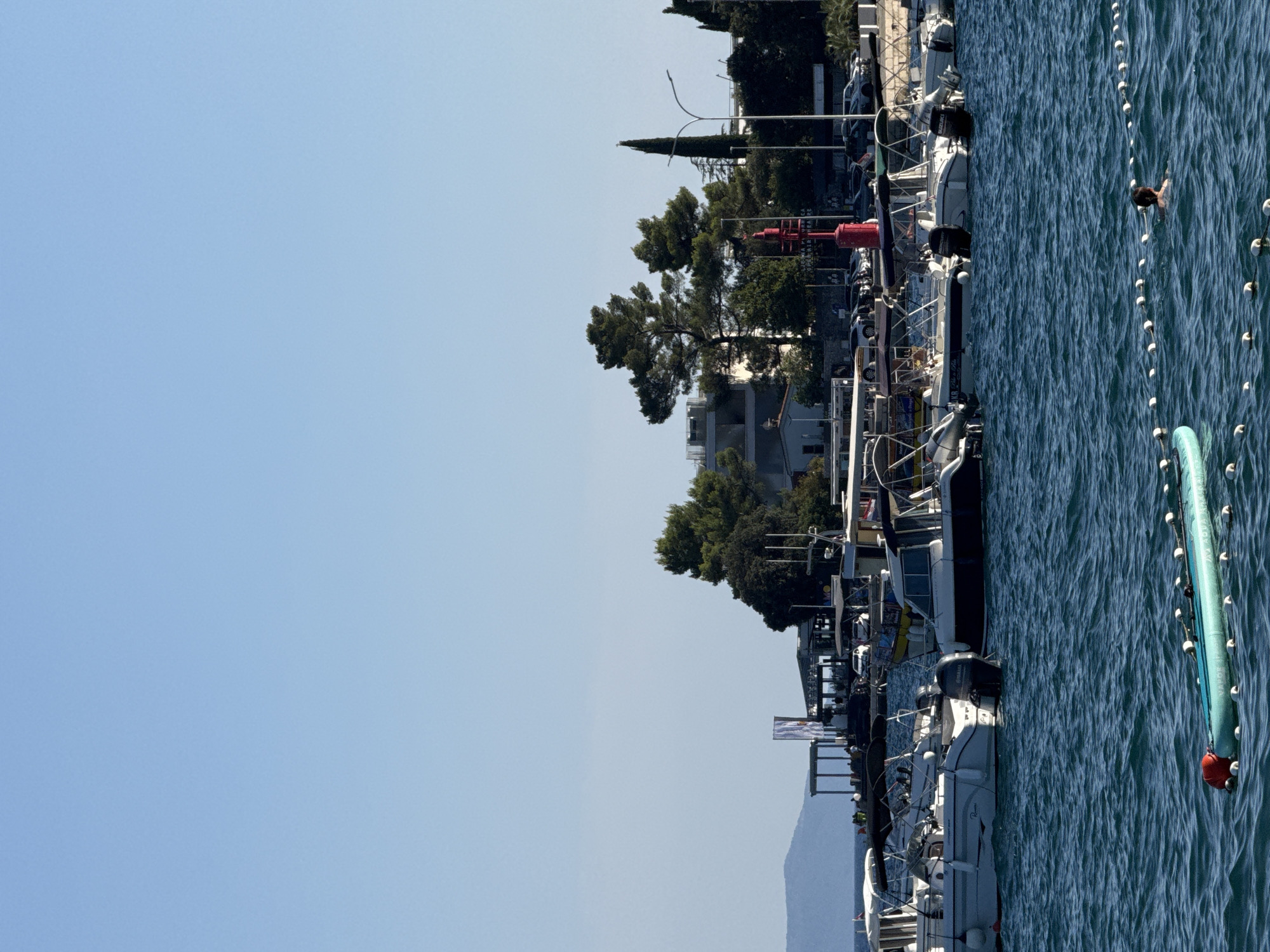 The image depicts a bright, sunny day in Grad Opatija, Croatia, overlooking a harbor. In the foreground, a person is swimming in the blue, slightly choppy water, with a rope barrier and buoys demarcating a swimming area. To the left, a pale blue paddleboard with a red buoy is visible. In the middle ground, several motorboats are docked in the harbor, with their white hulls and dark canopies reflecting the sunlight. Behind the boats, a picturesque coastline is lined with trees and buildings. A prominent red lighthouse stands near the shore, and further in the distance, a hazy mountain range is silhouetted against the clear blue sky. The scene is tranquil and evokes a sense of leisure and coastal charm.