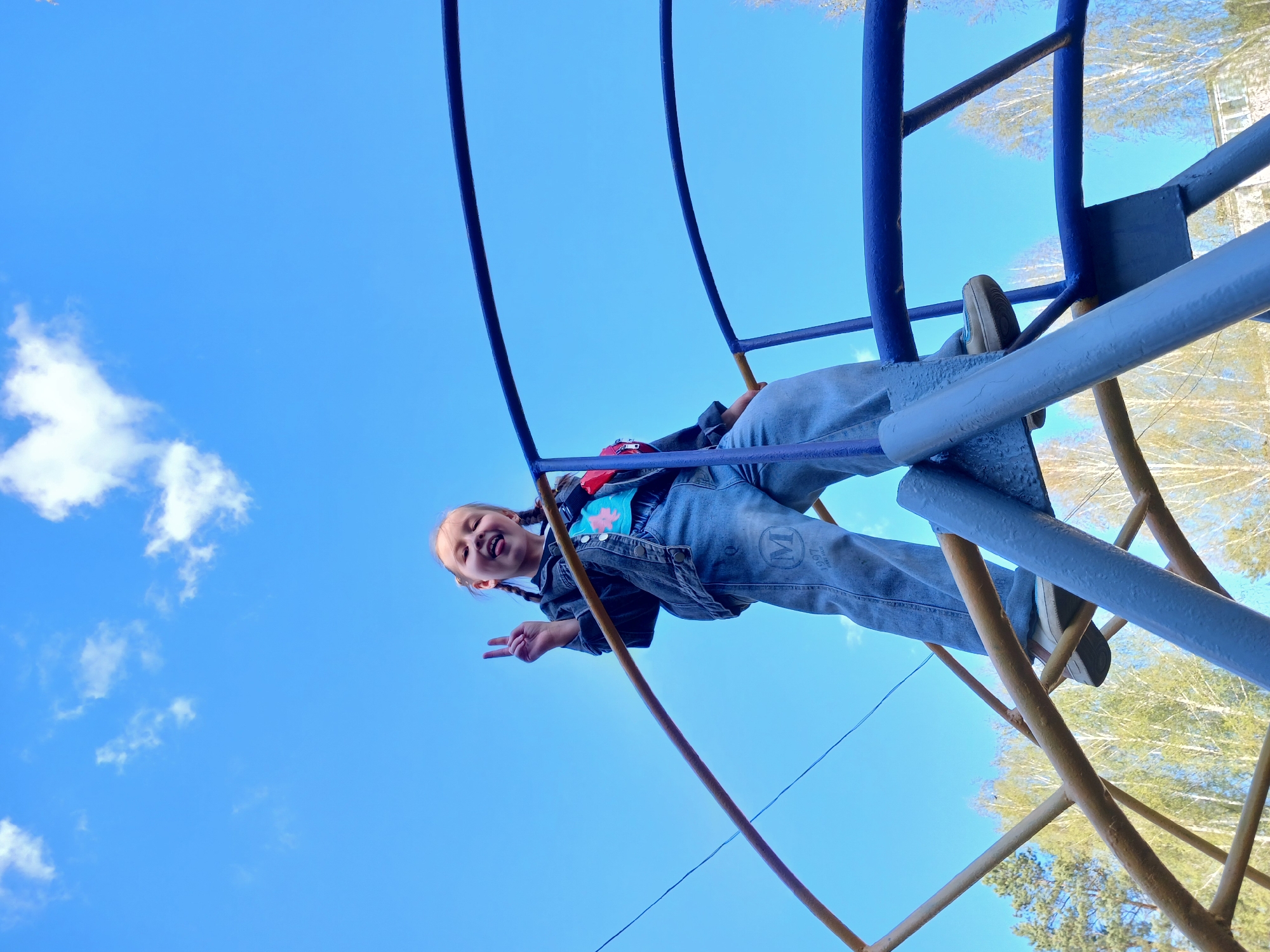 The image shows a girl on a playground structure against a backdrop of blue sky with scattered clouds. She is wearing a denim jacket, blue jeans with a visible "M" design, a T-shirt, and sneakers. She is positioned on the playground apparatus, possibly climbing or playing on it, as indicated by her placement among the blue and yellow bars and supports. Her expression suggests she is enjoying herself; she's smiling and making a peace sign with her fingers. The camera angle is low, looking up at her and the playground structure, emphasizing the height and the expansive sky. In the background, there are trees, suggesting an outdoor setting within a park or recreational area. The image was taken in the city of Noviy.