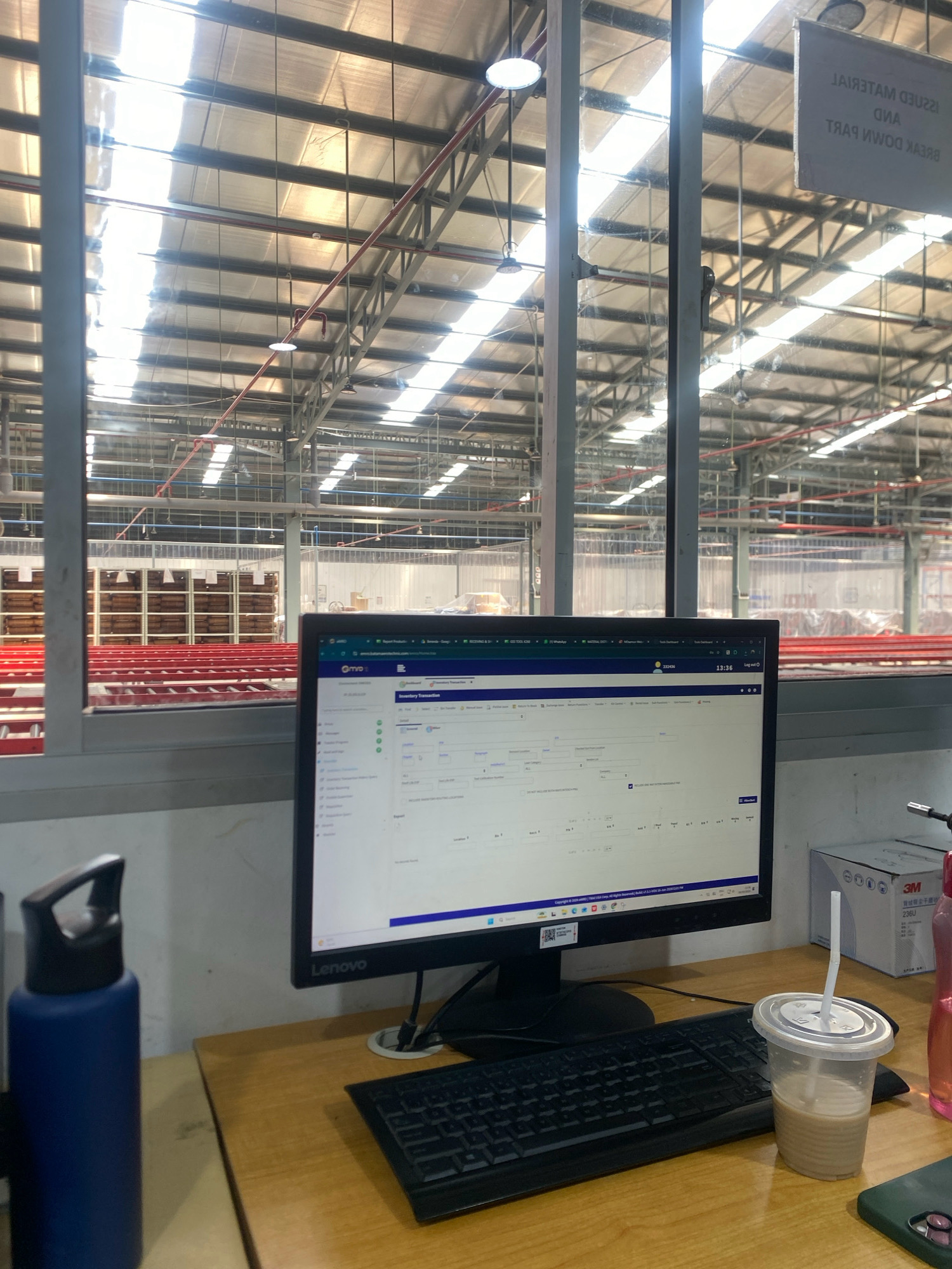 The image depicts a workstation set up within a large warehouse. The workstation features a Lenovo computer monitor displaying a software interface, a keyboard, and a mouse. On the desk, there's a blue water bottle and a clear plastic cup with a beverage and straw. In the background, through a glass partition, the expansive warehouse interior is visible, characterized by high ceilings with metal trusses and skylights, rows of shelving stocked with goods, and red conveyor belts. A sign with text in English and what appears to be an Asian script hangs in the upper right portion of the frame, visible through the glass. The overall environment suggests an operational logistics or distribution center. The image does not contain any discernible people, actions, or interactions.