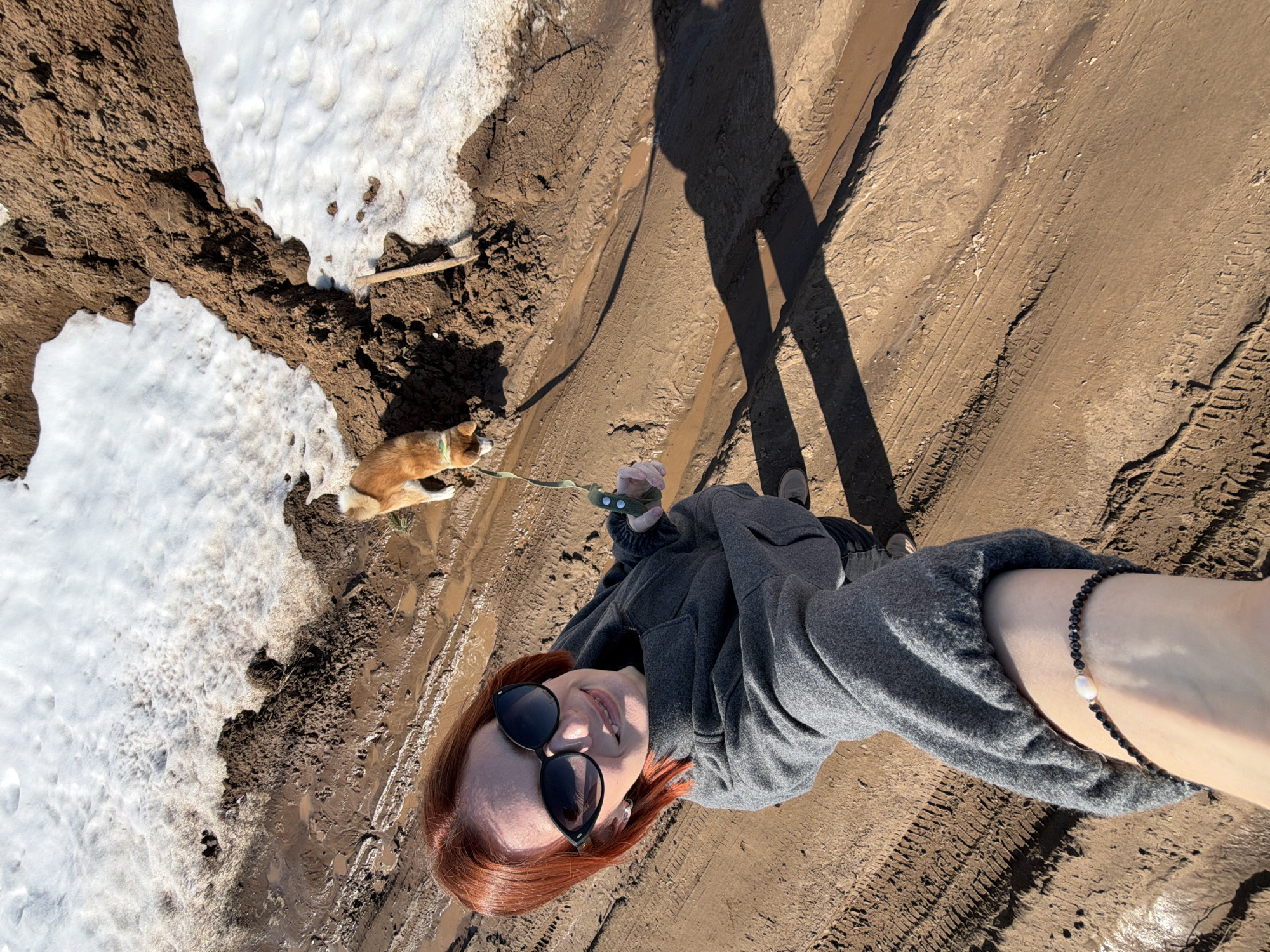 A selfie taken by a woman with red hair and sunglasses standing on a muddy path alongside a patch of snow and a small dog. The path shows tire tracks, suggesting vehicular traffic. The location is possibly in or near Chaikovsky, Russia. The woman is wearing a grey hoodie and a bracelet with a pearl. The dog is on a leash. A shadow of the woman is visible on the path.
