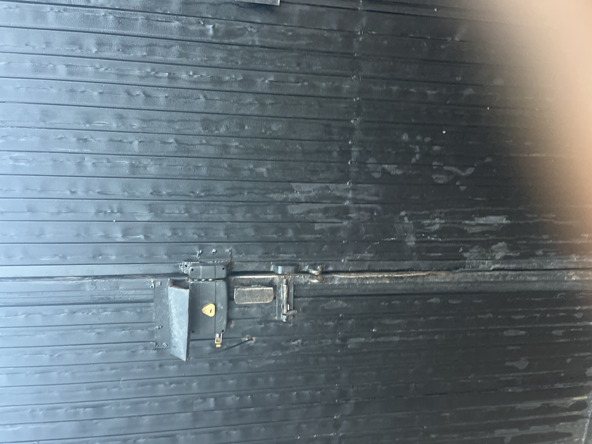 The image shows a close-up of a black, ribbed metal gate with multiple locks. The gate is weathered, with visible streaks and patches of lighter color suggesting wear and tear or possibly dirt. A prominent lock assembly is mounted on the left side of the gate, featuring a keyhole and a lever. Below this, another latch mechanism is visible, which appears to be engaged. The overall impression is of a functional, but aged security barrier. The image lacks any specific visual cues to pinpoint its location within Jos, Nigeria, such as landmarks, street signs, or distinctive architectural features.