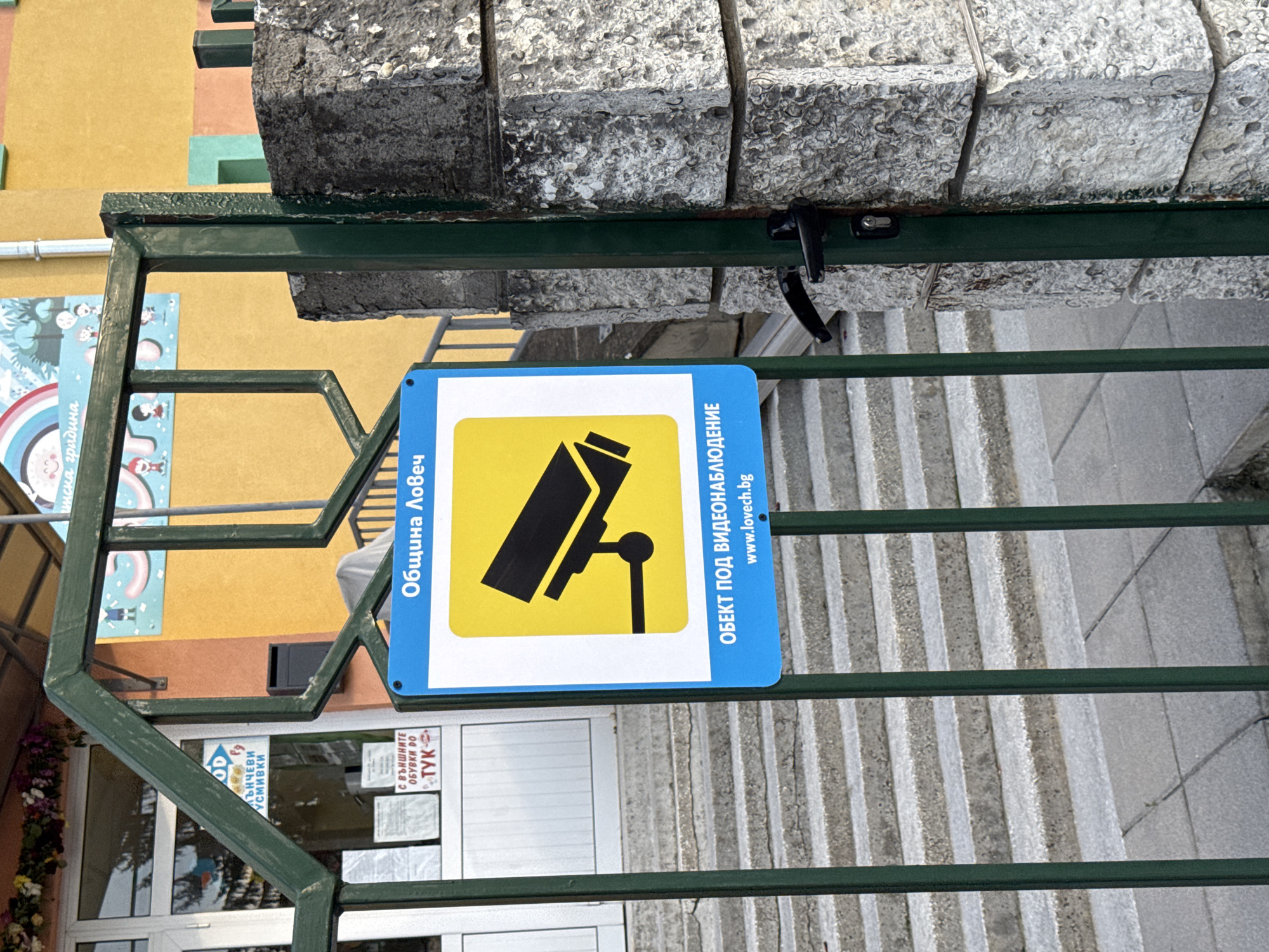 The image shows a green metal gate with a blue and yellow sign that reads "Община Ловеч" and "ОБЕКТ ПОД ВИДЕОНАБЛЮДЕНИЕ www.lovech.bg", which translates to "Municipality of Lovech" and "Object under video surveillance www.lovech.bg" in English. The sign features a stylized image of a surveillance camera. Behind the gate are concrete steps and a building with a sign that says "ЛЪНЧЕВИ УСМИВКИ" and "С ВЪНШНИТЕ ОБУВКИ ДО ТУК," which translates to "Lunchtime Smiles" and "With outdoor shoes to here." The background consists of a yellow and orange building facade. The location is in the city of Lovech, Bulgaria, based on the sign's text.