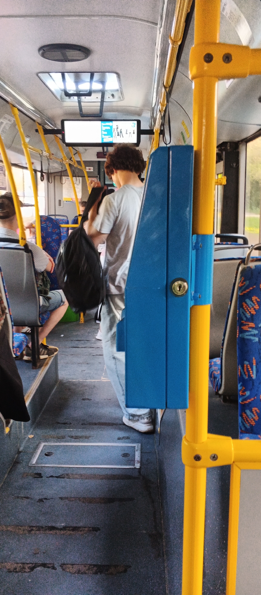 The image shows the interior of a bus in Izhevsk, Russia. A young man with curly brown hair, wearing a light grey t-shirt and light blue jeans, is standing in the aisle, holding a black backpack. He is facing away from the camera. To the left of the man, a passenger is seated, facing away from the camera. The bus interior features blue and orange patterned seats, yellow handrails, and a blue electronic device mounted on a yellow pole, which appears to be a ticket validator or information screen. A digital display screen is visible above the seats, showing some icons and text. The floor of the bus is dark grey with visible scuff marks and some metal grates. The overall lighting suggests it is daytime.