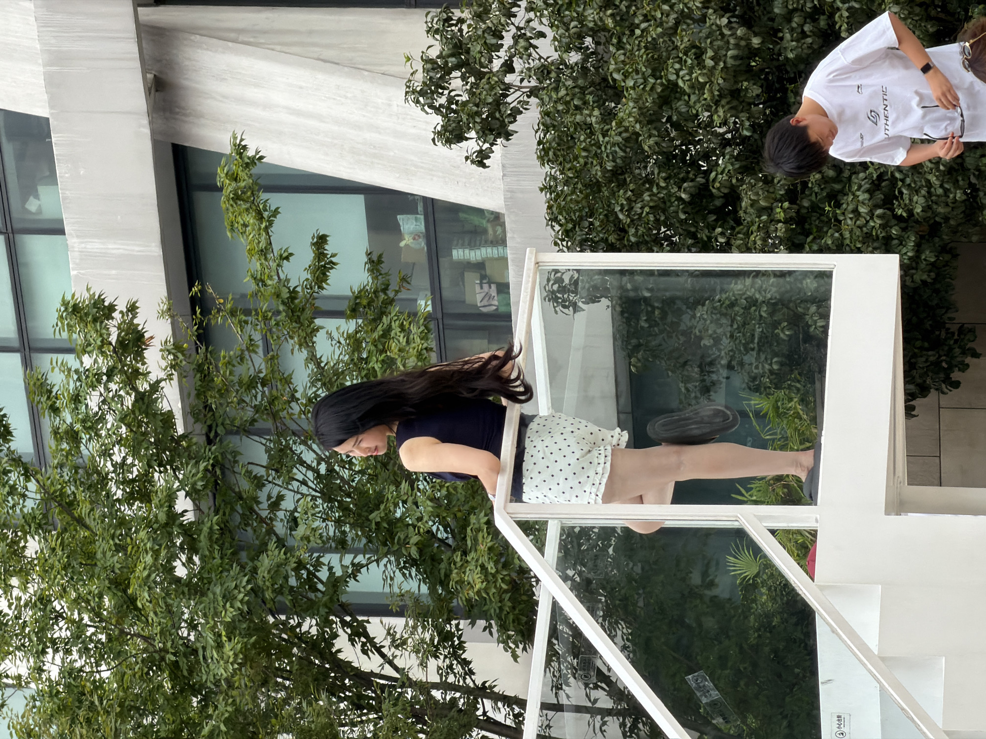 A young woman with long, dark hair is standing on a white platform with a glass railing. She is wearing a navy blue sleeveless top and white shorts with black polka dots. She is looking to her left, with her right hand resting on the railing. In the background, there are green trees and a modern building with large windows. Below the platform, a young man wearing a white t-shirt and black shorts is standing and looking down at something in his hands. He has short, dark hair and is wearing a black watch on his left wrist. The scene appears to be in an outdoor urban setting.