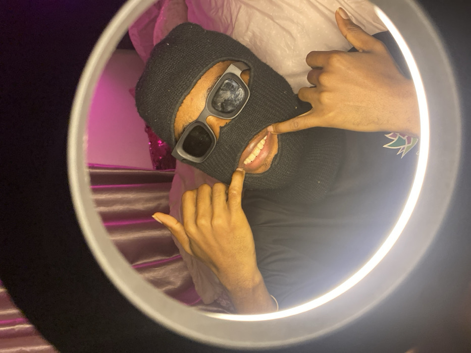 A person wearing a black ski mask and blocky sunglasses poses inside a ring light. Their fingers pull at the mask to expose their teeth. A glimpse of their patterned shirt is visible near their shoulder. The background shows pink lighting, shiny fabric, and a pillow.