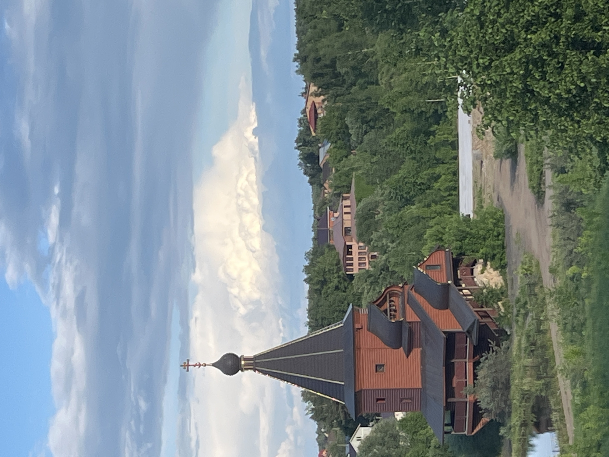 The image features a wooden church with a traditional Russian Orthodox design, topped with a black onion dome and a cross, located in Большое Саврасово, Russia. It is situated amongst lush green trees and buildings. The church is built on a slope. A dirt path can be seen running past the church on the right. The sky is blue with scattered clouds.
