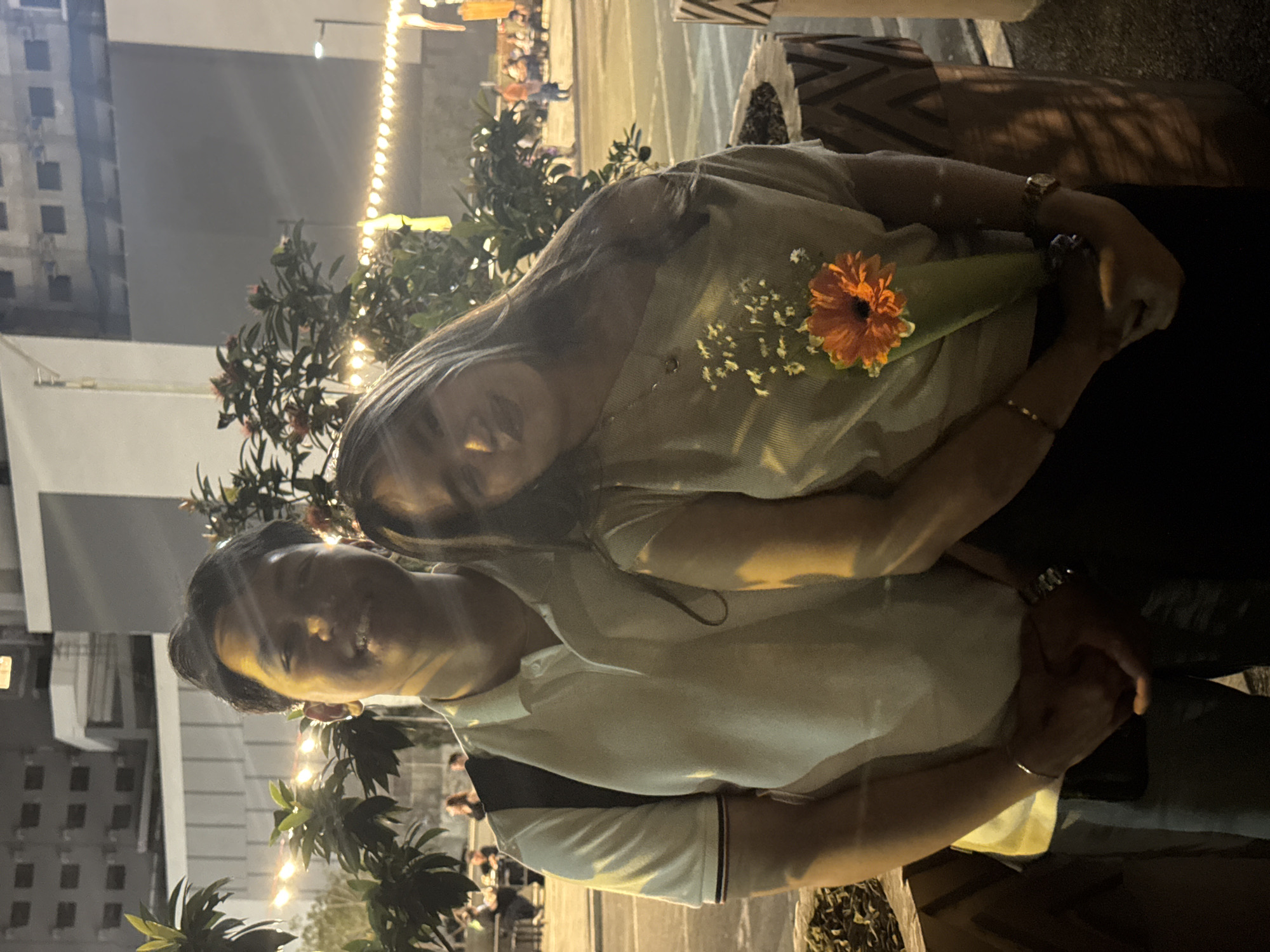 Two people are posing for a photo at night, with a background suggesting an outdoor event or gathering. The person on the left has short, dark hair and is wearing a light-colored shirt with dark trim on the shoulder. The person on the right has long hair and is holding a flower. Lights and greenery are visible in the background. A building is also visible in the background. There are multiple other people in the background. The location is likely Davao City, Philippines.