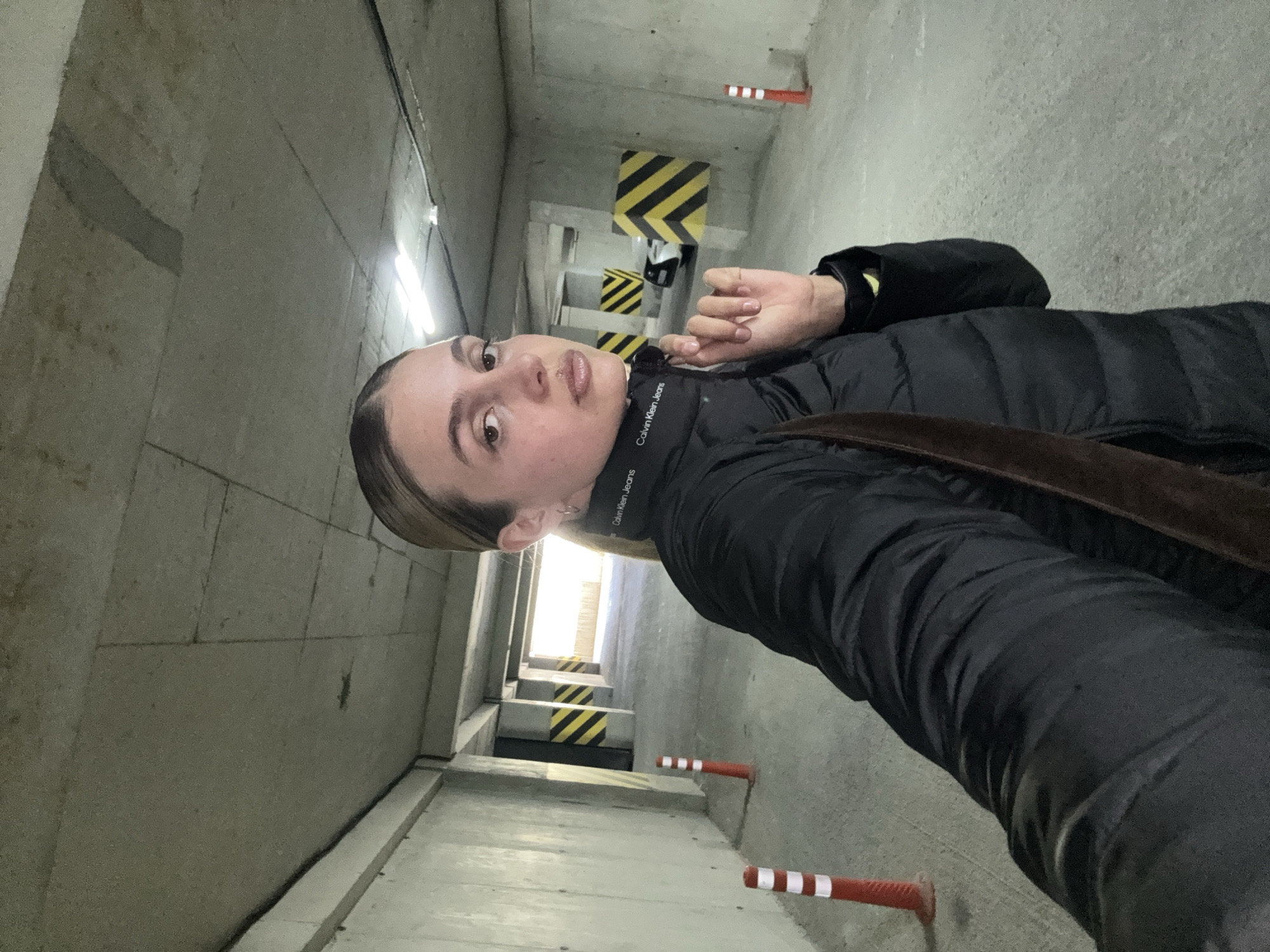 A woman is taking a selfie in what appears to be a parking garage or similar industrial setting. The location, Unknown, Unknown, is implied by the absence of any information about location. The woman is wearing a black puffer jacket with a Calvin Klein Jeans collar visible. The walls around her are concrete and the lighting is dim, likely from fluorescent lights in the ceiling. In the background, there are striped yellow and black barricades and red and white safety posts along the wall. The image is taken at an angle, making the scene appear tilted.