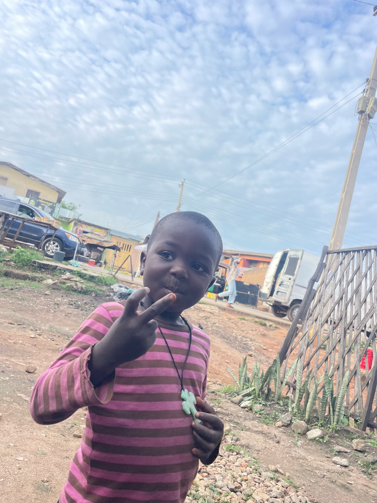 In Jos, Nigeria, a young child with dark skin is captured in a medium shot, posing with their right hand raised, showing two fingers in a peace sign. The child is wearing a long-sleeved pink and brown striped shirt and a light green pendant shaped like a frog on a black string around their neck. They have dark eyes, a small nose, and their mouth is slightly open, revealing teeth. The background depicts an outdoor scene with a dirt road, scattered rocks, and sparse vegetation, including a row of snake plants. In the distance, there are buildings, a parked blue SUV, and a white van. A power pole with electrical wires and a metal fence are also visible, contributing to the urban but somewhat unkempt environment. The sky is overcast with patchy clouds.