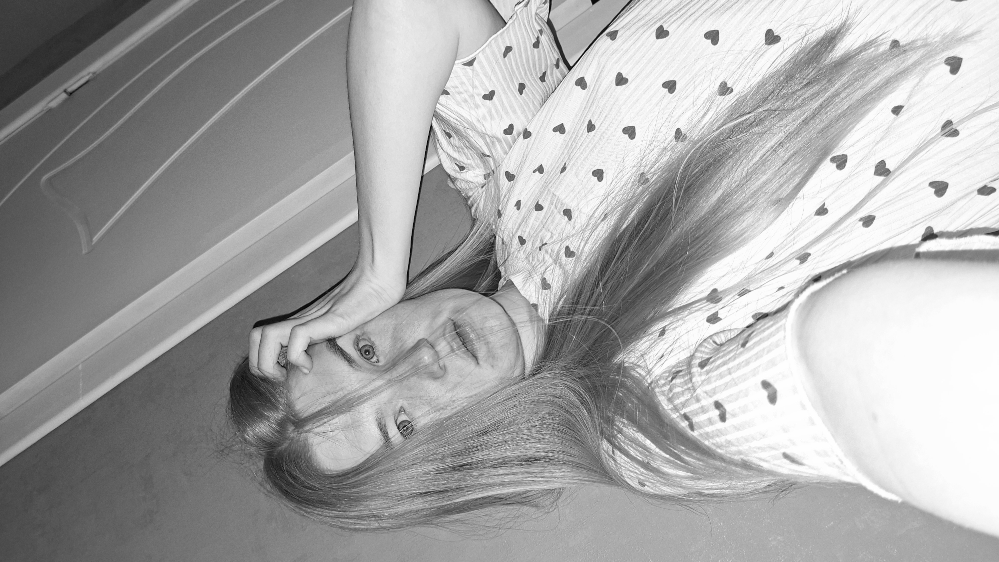 A grayscale close-up of a woman lying on her side, looking up at the camera. She is wearing pajamas with hearts. Her long hair is spread out around her. One hand is near her face, possibly touching her forehead or hair. A door frame or edge of a wall is visible in the background. The location in the city of Chaikovsky, Russia, is not discernible.