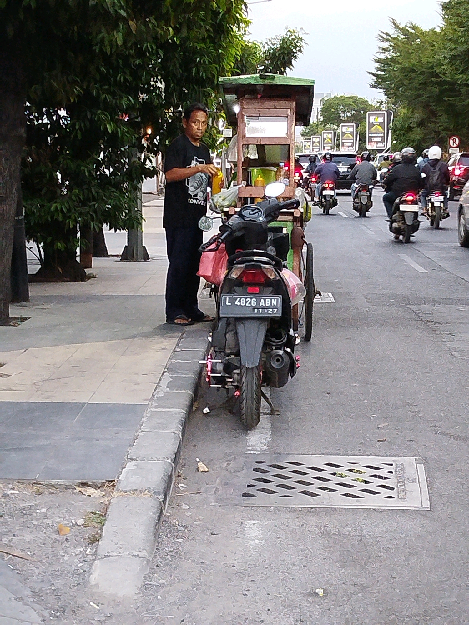 A street scene in Surabaya, Indonesia, features a street vendor standing next to a motorcycle with a sidecar, which is loaded with goods. The vendor, a man wearing a black t-shirt and dark pants, holds a yellow object in his hand. The motorcycle, a black scooter with license plate "L 4826 ABN", has a red taillight and a silver exhaust pipe. In the background, a busy street with traffic, including other motorcycles and cars, is visible. The street is lined with trees and buildings. The ground is paved with concrete and asphalt.