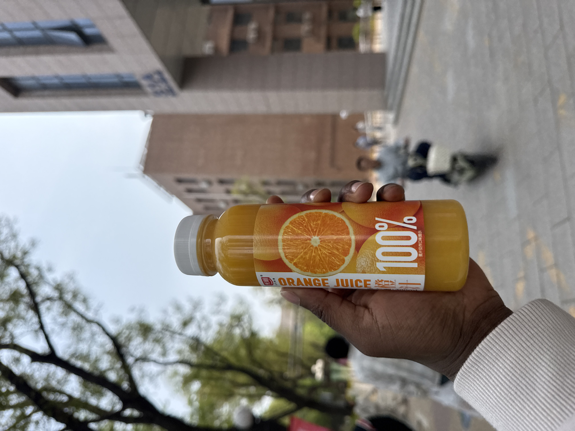 A bottle of orange juice is held up in a person's hand in front of a blurred city scene. The orange juice bottle is a plastic cylindrical shape with a white cap. The label shows a cross-section of an orange and the words "100% ORANGE JUICE". The hand holding the bottle has dark skin, and the person wears a white long-sleeved shirt. In the background, there is a blurred scene of a modern building and trees, suggesting an urban environment. The photograph was taken in Daxing District, China.