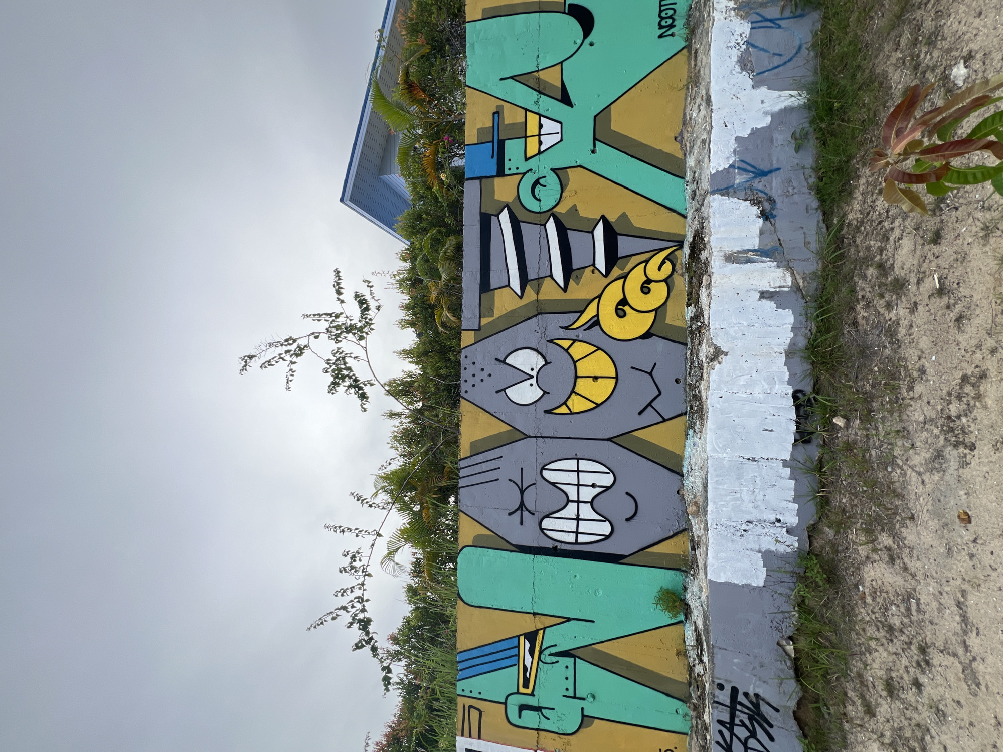 An outdoor, eye-level, wide shot captures graffiti art on a low, concrete wall in Ko Phangan, Thailand. The graffiti features cartoonish figures forming letters or words, primarily in green and gray with yellow accents. The top portion of the image shows a cloudy sky with some green foliage and a building with a blue roof in the background. The bottom portion of the image depicts the ground in front of the wall, composed of dirt and sparse vegetation. The concrete wall has a rough, weathered texture, with areas of peeling paint. The graffiti depicts at least three distinct characters: one on the far left with a green body, blue accents, and a yellow element near its mouth; two gray characters with exaggerated facial features, one with angry eyes and the other with a wide smile; and a final green character on the far right wearing a blue hat. The overall atmosphere is artistic and somewhat urban, despite the natural elements present.