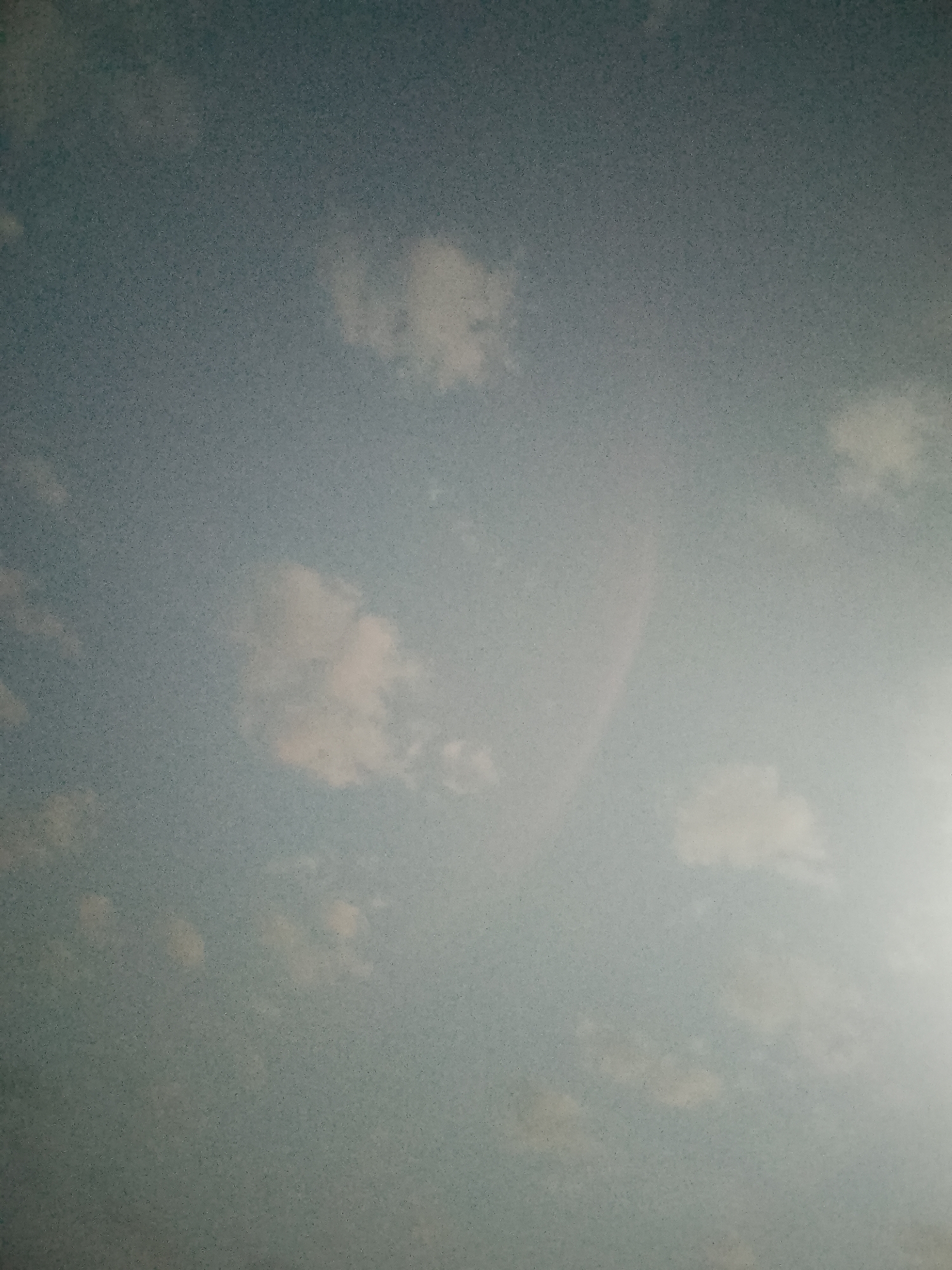 The image depicts a pale blue sky with scattered white clouds. A faint, rainbow-like arc is visible in the center-right area of the image. The location appears to be outdoors, with no indication of a city or specific geographical location.