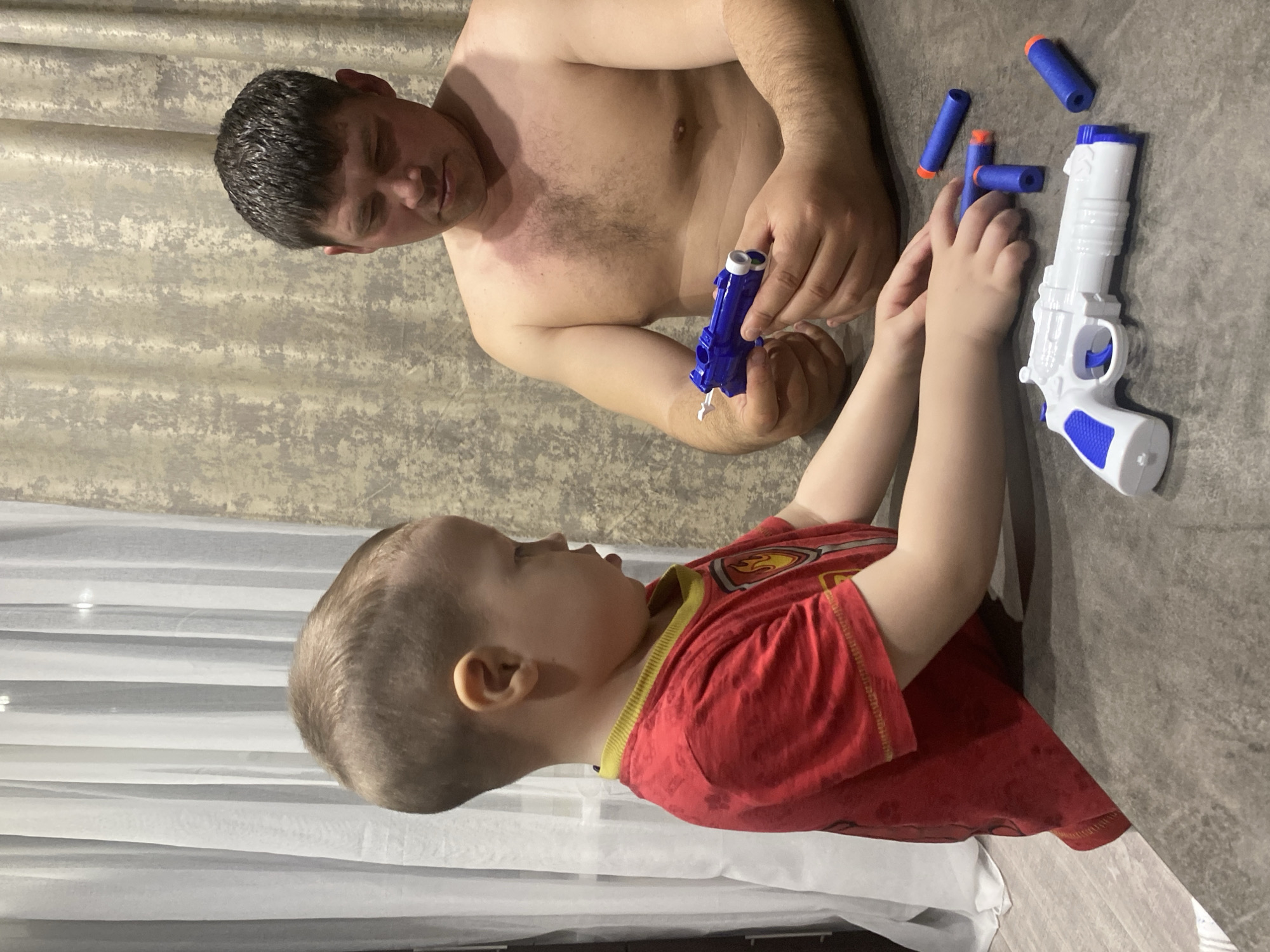 The scene depicts two individuals, an adult male and a young child, at a dark grey, textured table in an indoor setting located in the city of Sochi, Russia.
The adult male, shirtless with a visible tan and a reddish patch on his upper left chest, is seated on the right side of the frame. He has short, dark, slightly wet-looking hair and appears to be focused downwards, examining a blue toy pistol-like object held in both hands. The toy has white or light-colored tips at the barrel end.
On the left, a young child, approximately three to five years old, stands with his back partially to the camera, facing the table. He has short, light brown hair and wears a red short-sleeved t-shirt with a yellow ribbed collar and a yellow and red emblem on the chest. His hands are resting on the table near a white and blue toy revolver-like object.
Scattered on the table near the child's hands and the white toy gun are four blue foam darts with orange tips.
The background features two sets of curtains: a sheer white curtain on the left and a textured, light beige/grey patterned curtain on the right, suggesting a window or room divider behind the subjects. The floor is not visible. The lighting appears to be from an overhead source.