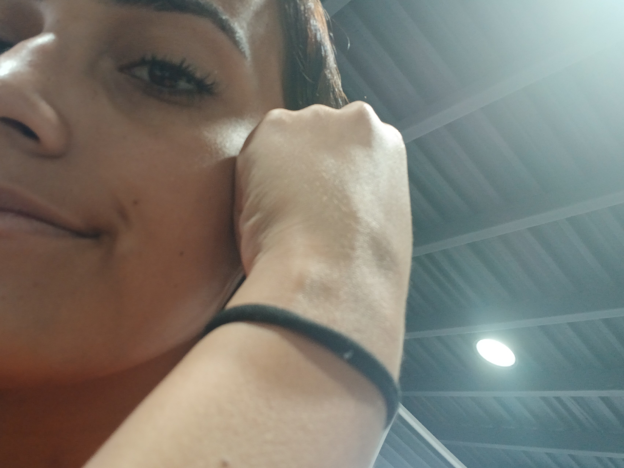 The image shows a close-up of a person's face and arm, with their hand resting near their cheek. The person has dark hair and is wearing a black elastic band on their wrist. The background appears to be an indoor ceiling with exposed beams and a light fixture. The angle of the shot is from below, looking up. Without additional information, the specific location in Juan Griego, Venezuela cannot be determined from the visual content of the image alone.