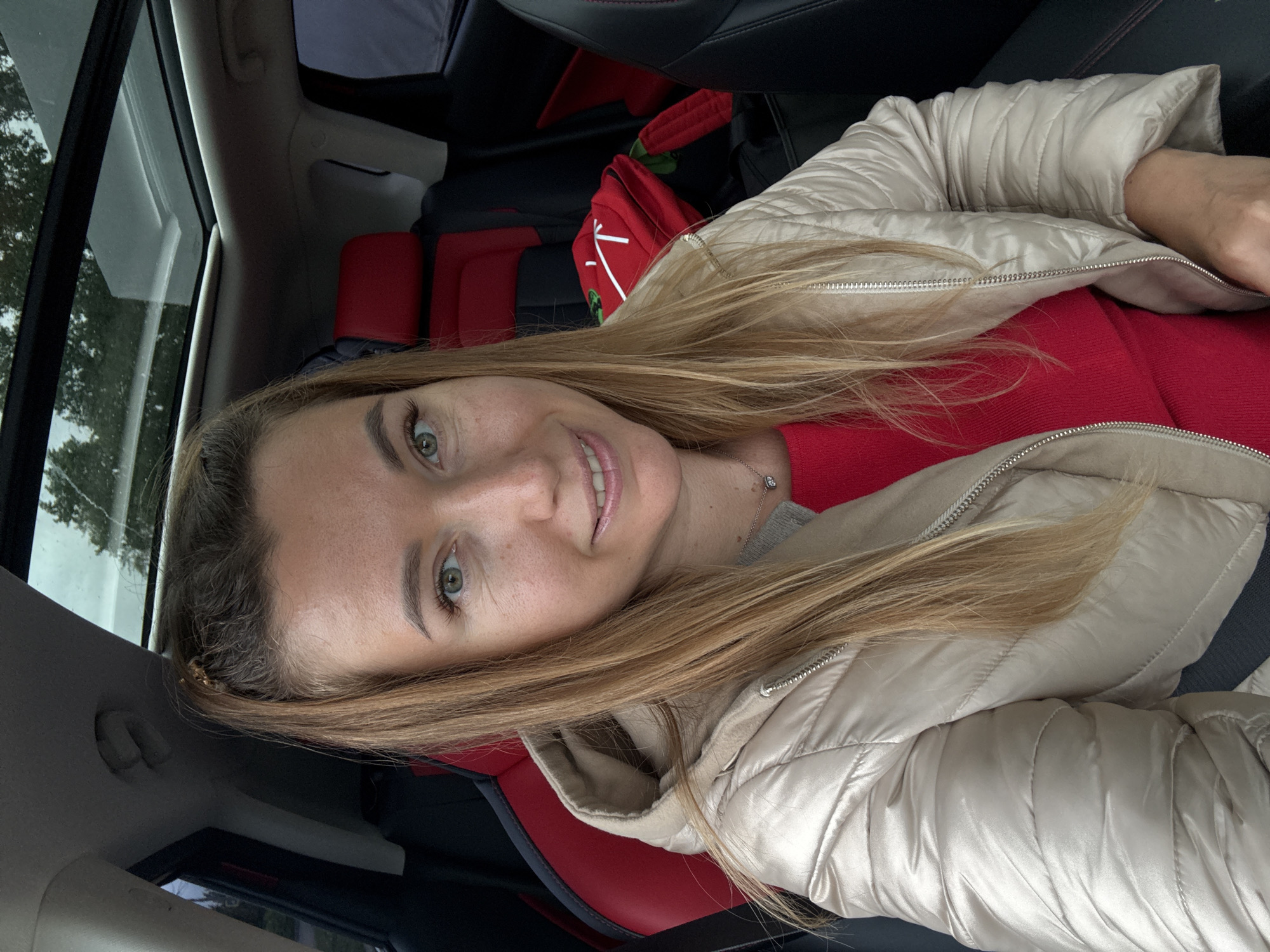 A woman is seated in a car, possibly a passenger. She is wearing a light beige puffer jacket over a red top. The car has red and black leather seats. The woman has blonde hair and is smiling. The car's interior is visible, including part of the windshield and sunroof. Outside, trees are visible through the window. The location is Unknown, Unknown.