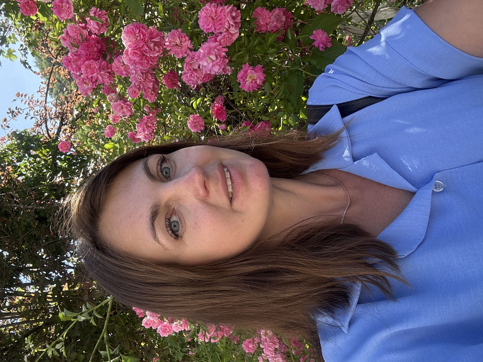 A medium shot, selfie-style photograph features a woman with long, brown hair and blue eyes, smiling slightly. She is wearing a light blue, collared shirt and a thin silver necklace. The background is dominated by a dense bush of bright pink roses in full bloom, with green foliage and branches visible. The lighting suggests it is a sunny day. There are no discernible landmarks indicating the specific location within Saint-Claude, France. The image captures a woman posing in front of a rose bush.