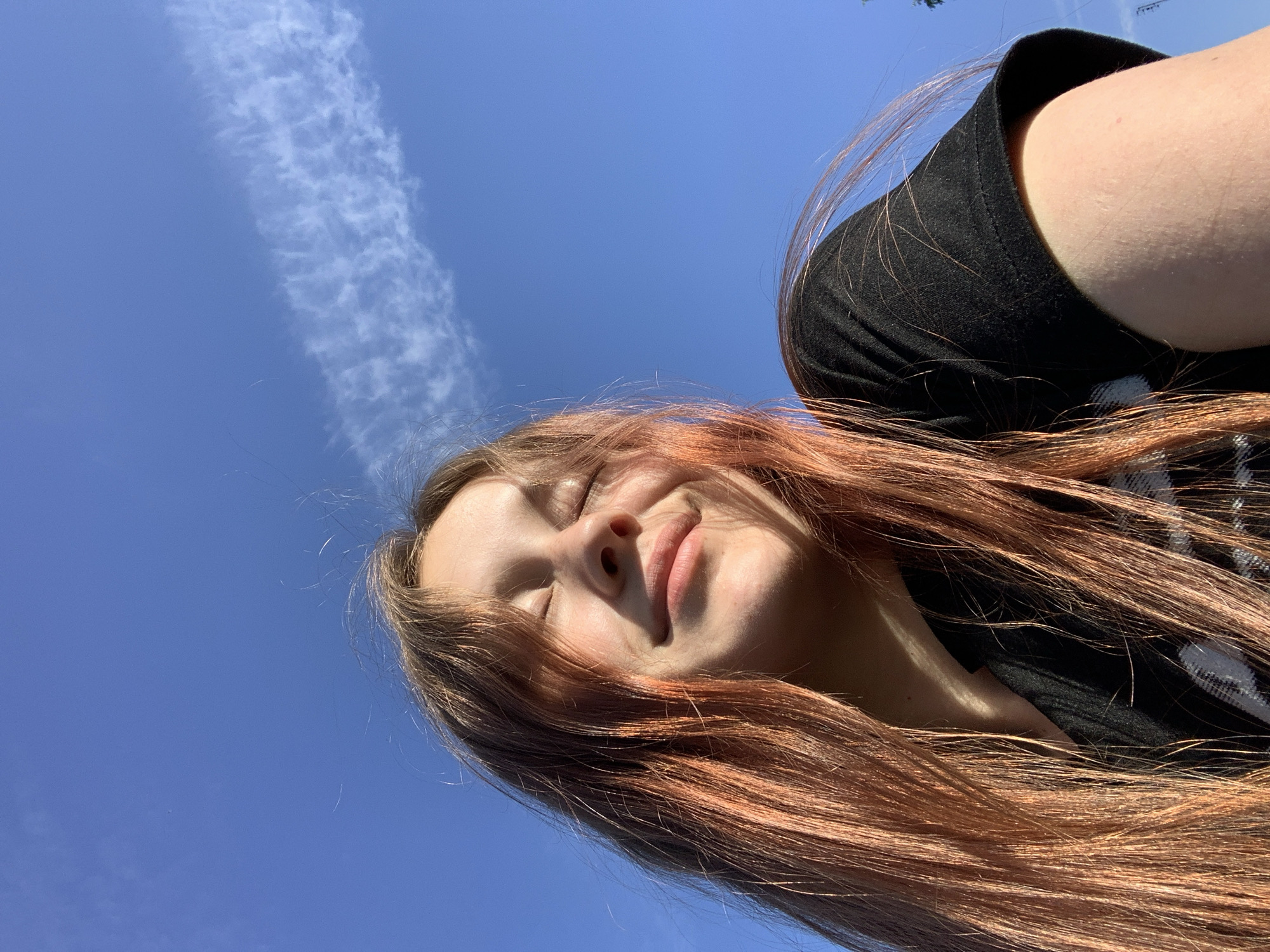 The image is a low-angle shot of a person against a bright blue sky with a wispy white cloud formation. The person has closed eyes, a slight smile, and long, reddish-brown hair that is partially illuminated by sunlight. They are wearing a black t-shirt. The location cannot be determined based solely on the image; therefore, the request for "Foki, Russia" cannot be fulfilled.