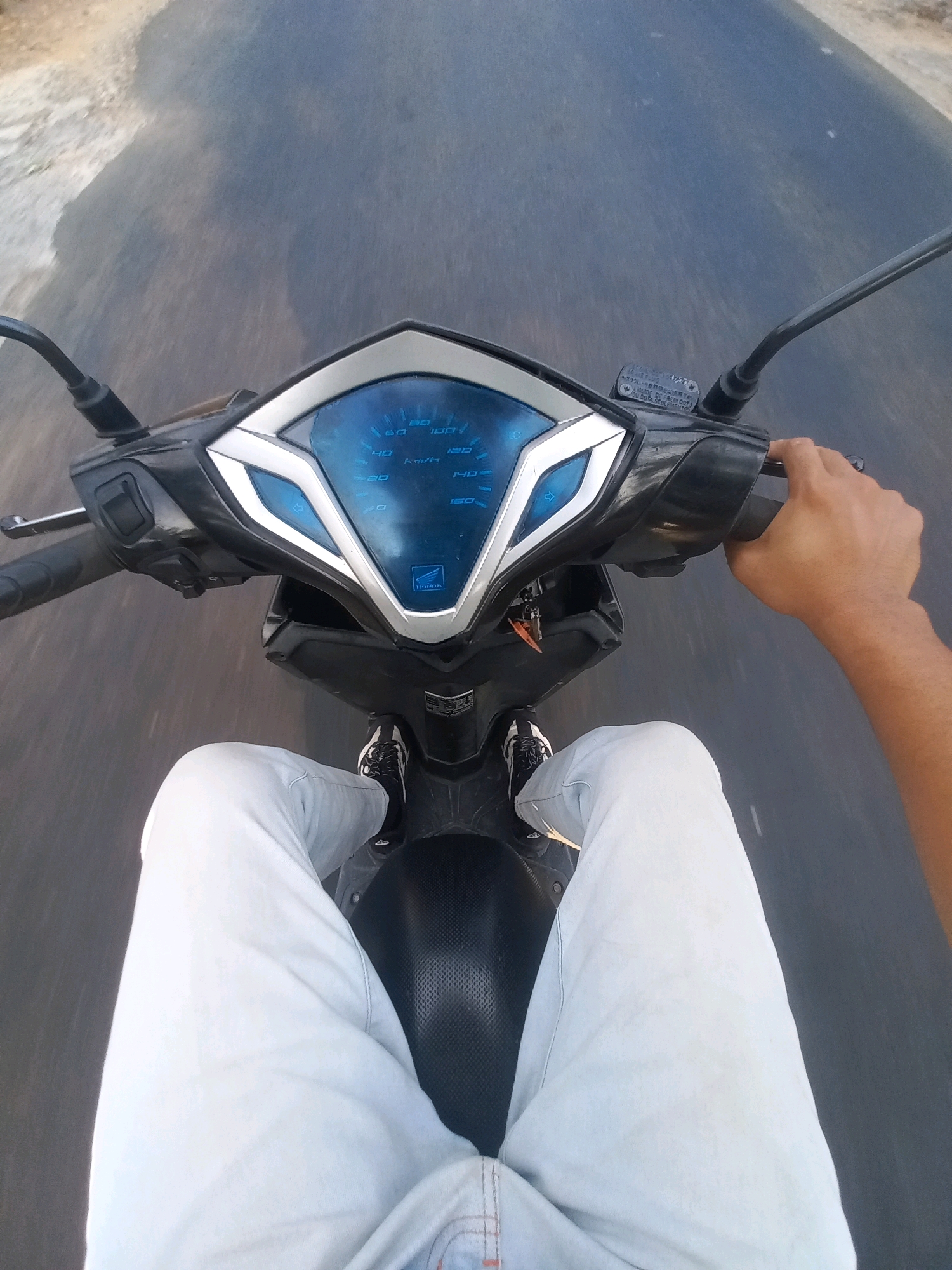 An individual is riding a scooter on a road. The viewpoint is from the rider's perspective, looking down at the handlebars and their legs. The scooter has a digital speedometer with blue accents. The rider is wearing light-colored pants and black and white sneakers. The road appears to be asphalt with some debris on the left side. The overall environment suggests an outdoor setting, possibly a street or highway. The context of Tanjungsari, Indonesia, indicates a tropical or subtropical climate. The image is a first-person perspective of someone in motion on a scooter.