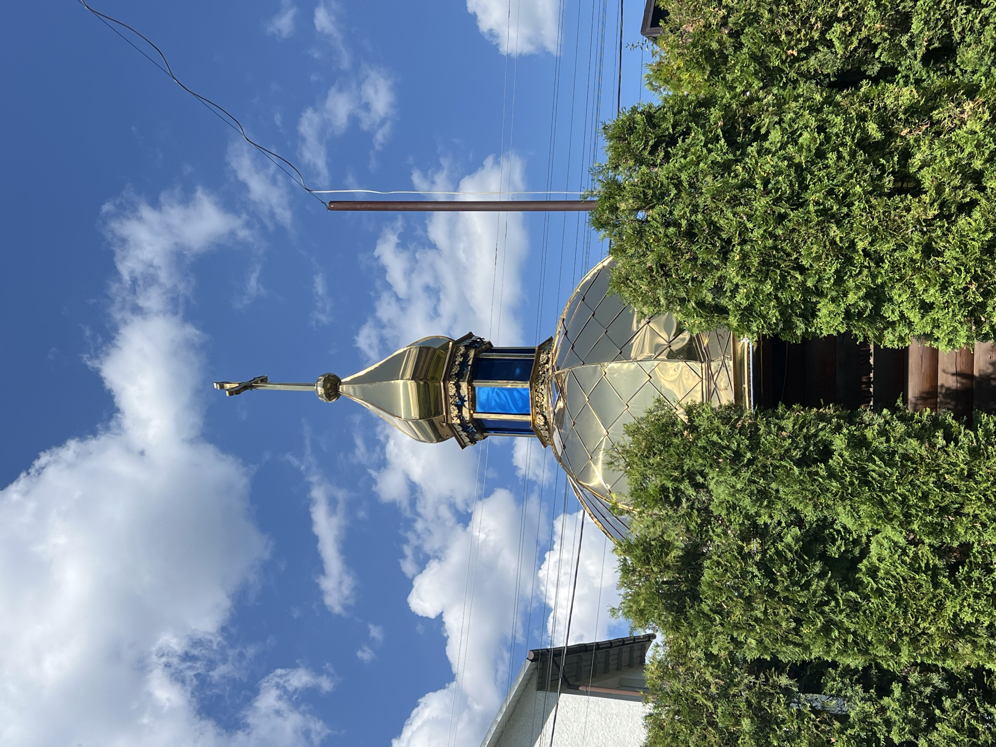 The image shows the golden dome of a church in Yaremche, Ukraine, partially obscured by lush green trees. The dome is topped with a cross and features blue architectural elements. In the background, a clear blue sky with scattered white clouds is visible, along with power lines and a tall, thin pole. To the left, a portion of a white building with a grey roof is visible. The overall scene suggests a sunny day in a picturesque, possibly rural, setting within the city.